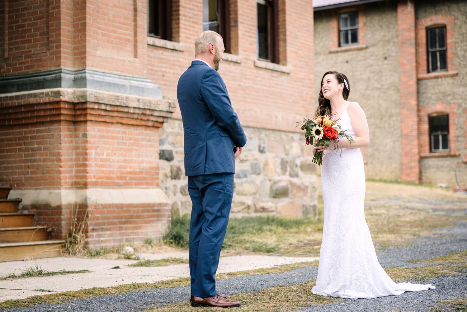 A couple in formal wedding attire poses together in front of a rustic brick building during their special day.