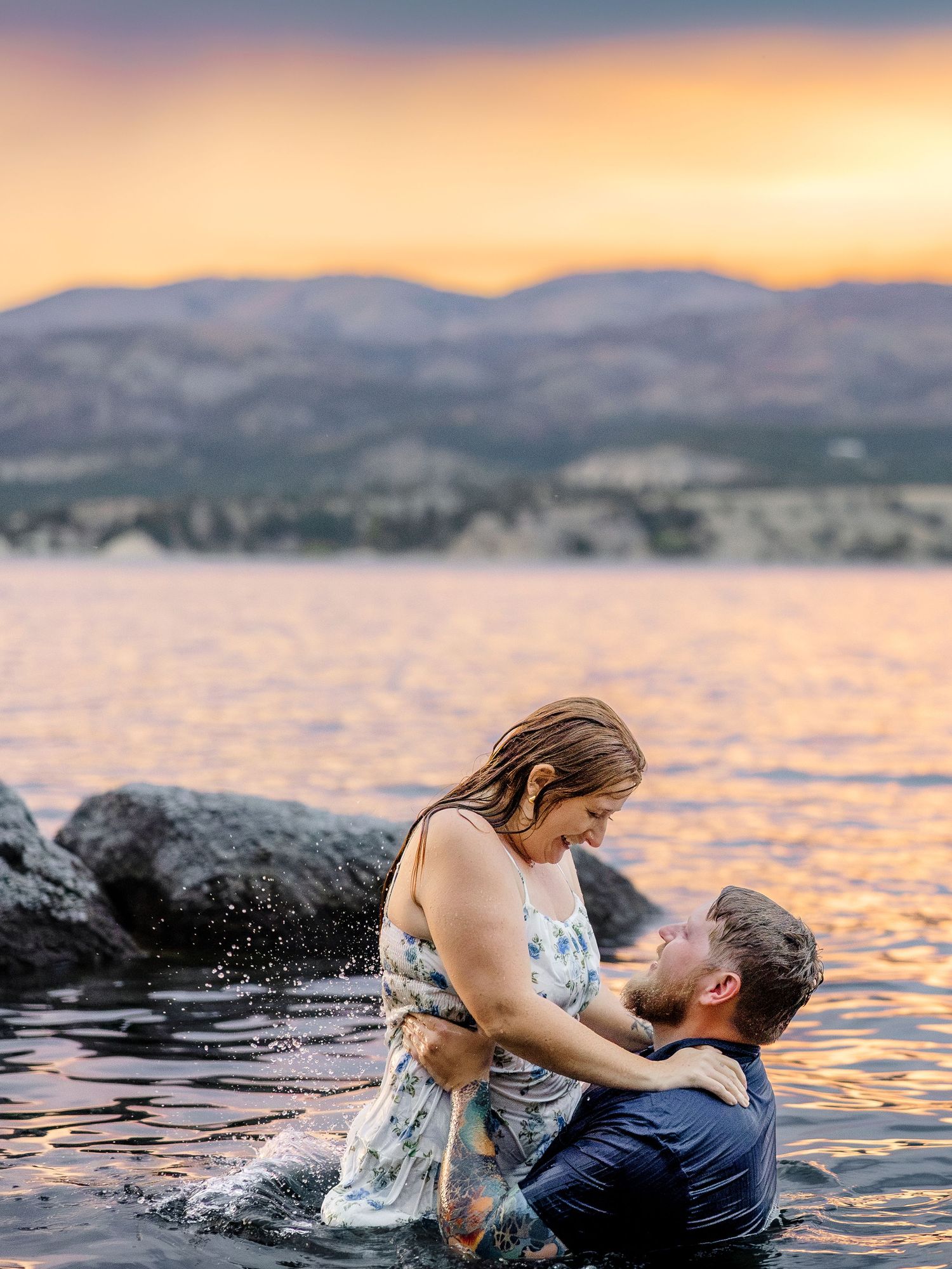 Couple embracing in a lake at sunset with golden skies and mountain backdrop during an engagement photo session.