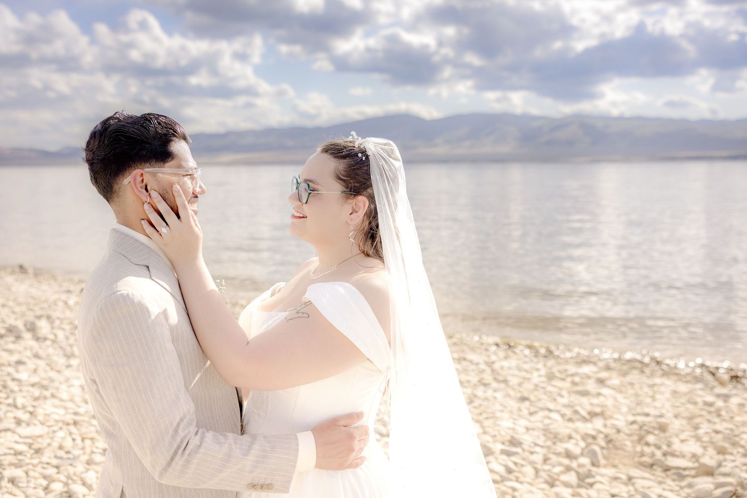 A bride and groom share a romantic moment on a sandy beach with mountains and cloudy sky in the background.