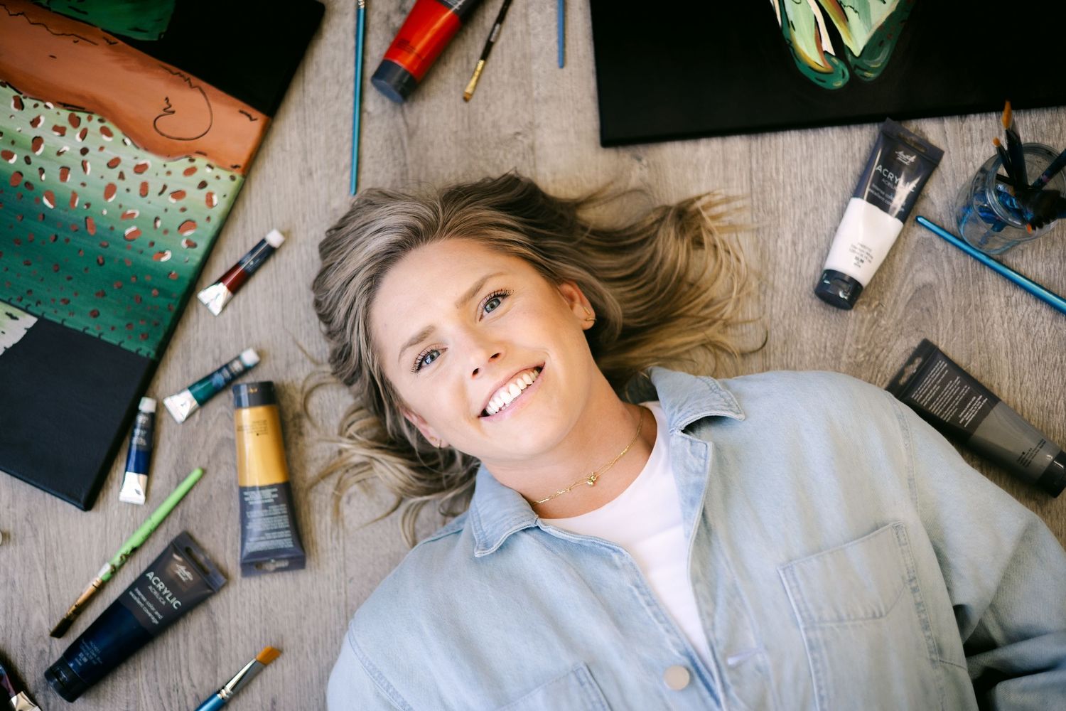 Smiling woman lying on wooden floor surrounded by art supplies, paint tubes, brushes, and sketchbooks from above.