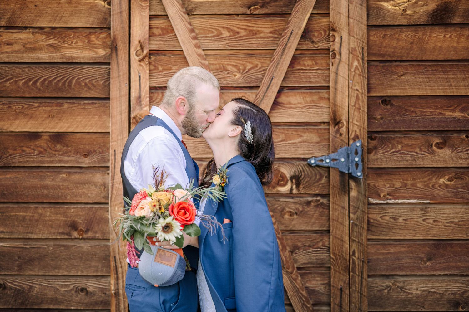 A couple shares a romantic kiss against a rustic wooden barn wall while holding a colorful autumn bouquet.