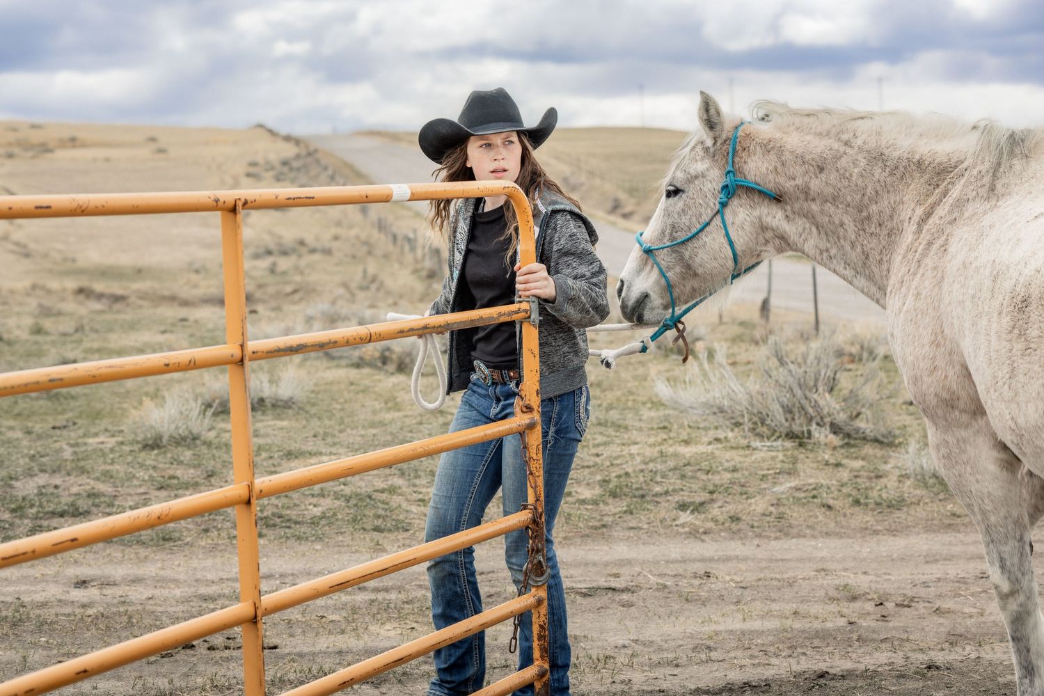 A Montana cowgirl in western attire leans opens an orange metal fence in a rural landscape while leading her horse during winter.