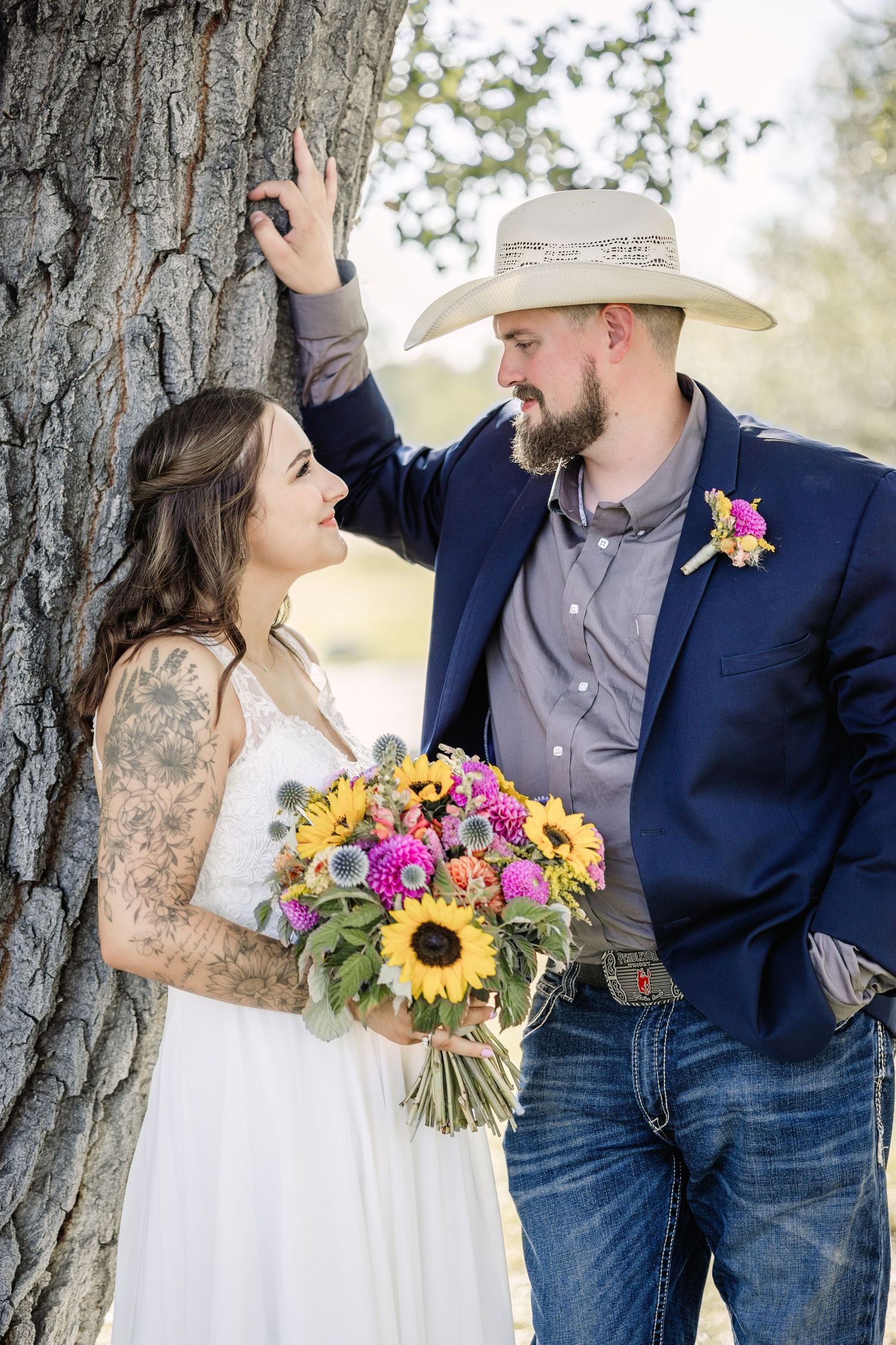 A Helena wedding couple poses by a tree trunk with a vibrant sunflower and wildflower bouquet.