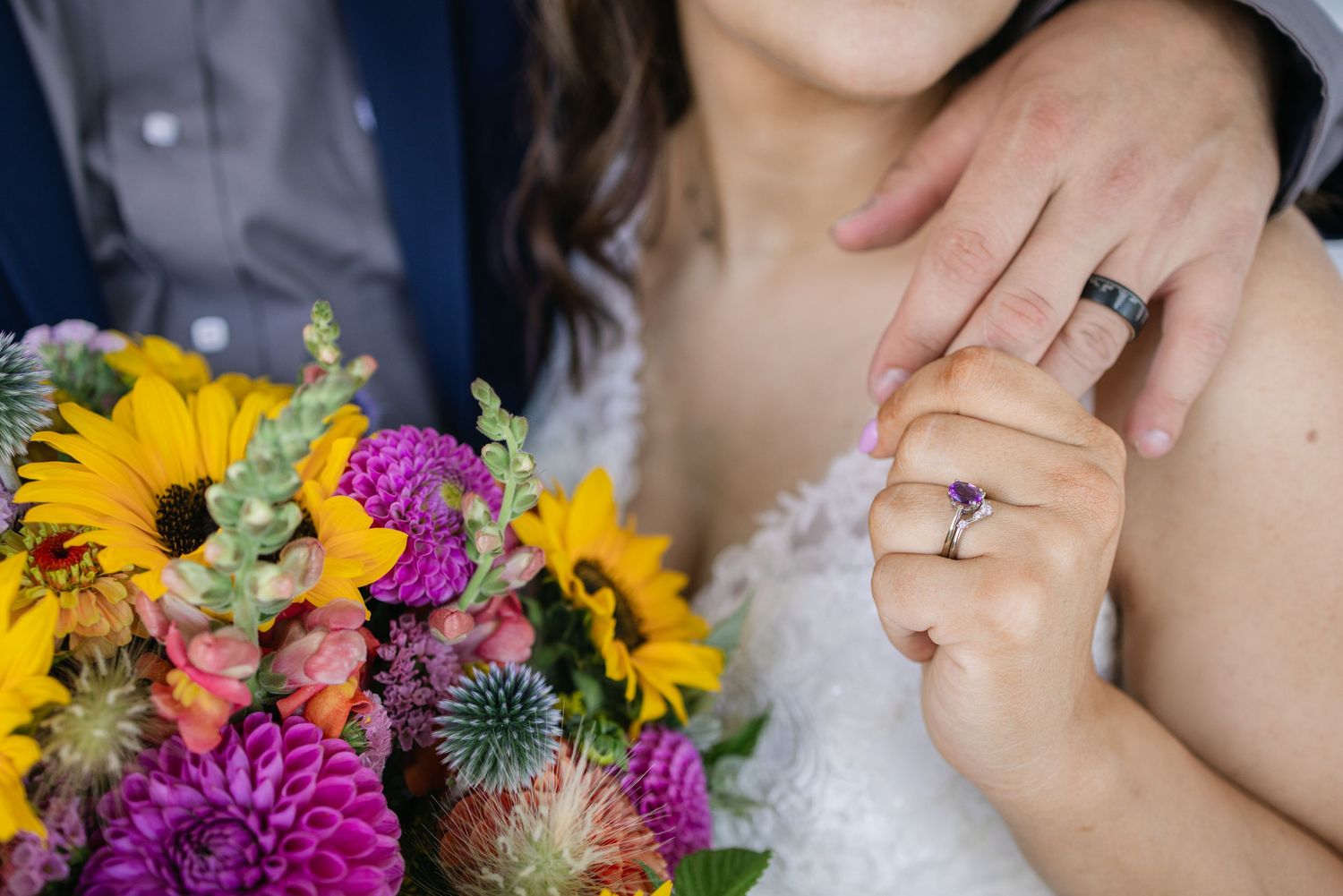 A hand wearing a wedding ring holds a vibrant bouquet of sunflowers and purple flowers.