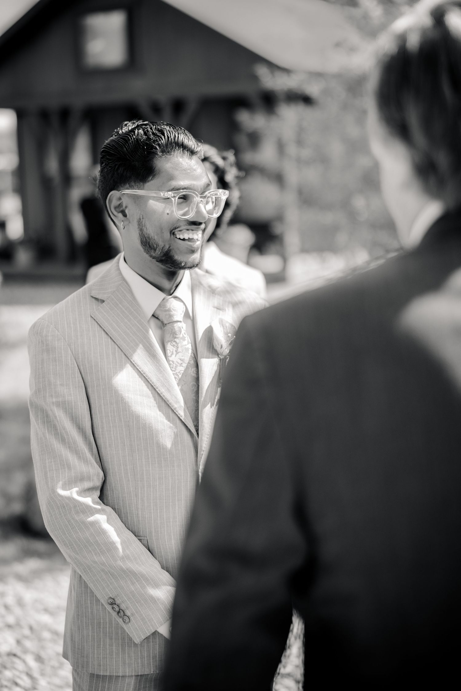 Black and white photo of a smiling groom in a pinstripe suit and glasses chatting with a guest at an outdoor wedding.