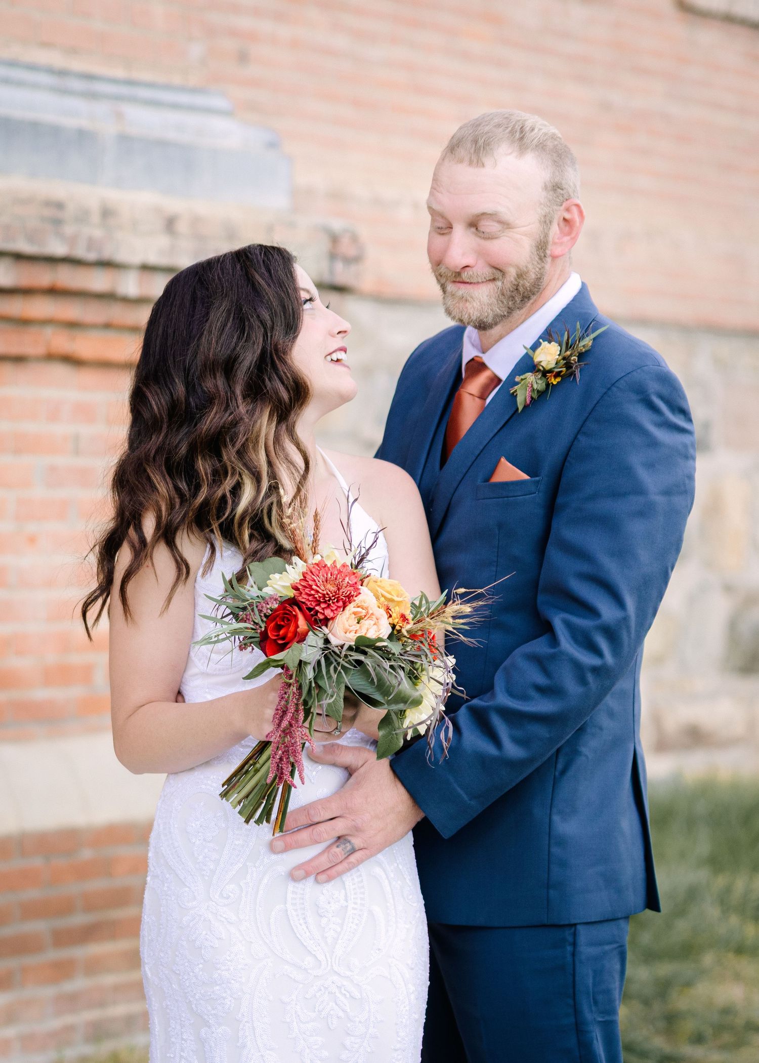 A couple in wedding attire share a joyful moment against a brick wall backdrop during their outdoor photo session.