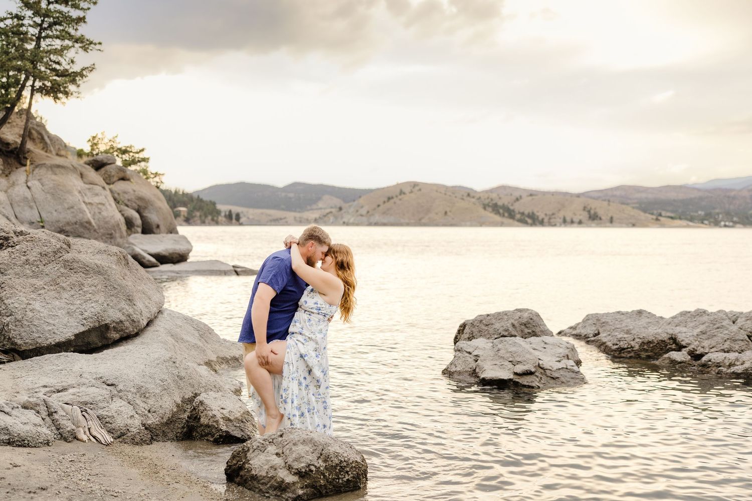 Couple sharing a romantic kiss on rocky shoreline with serene lake and mountain backdrop at golden hour.