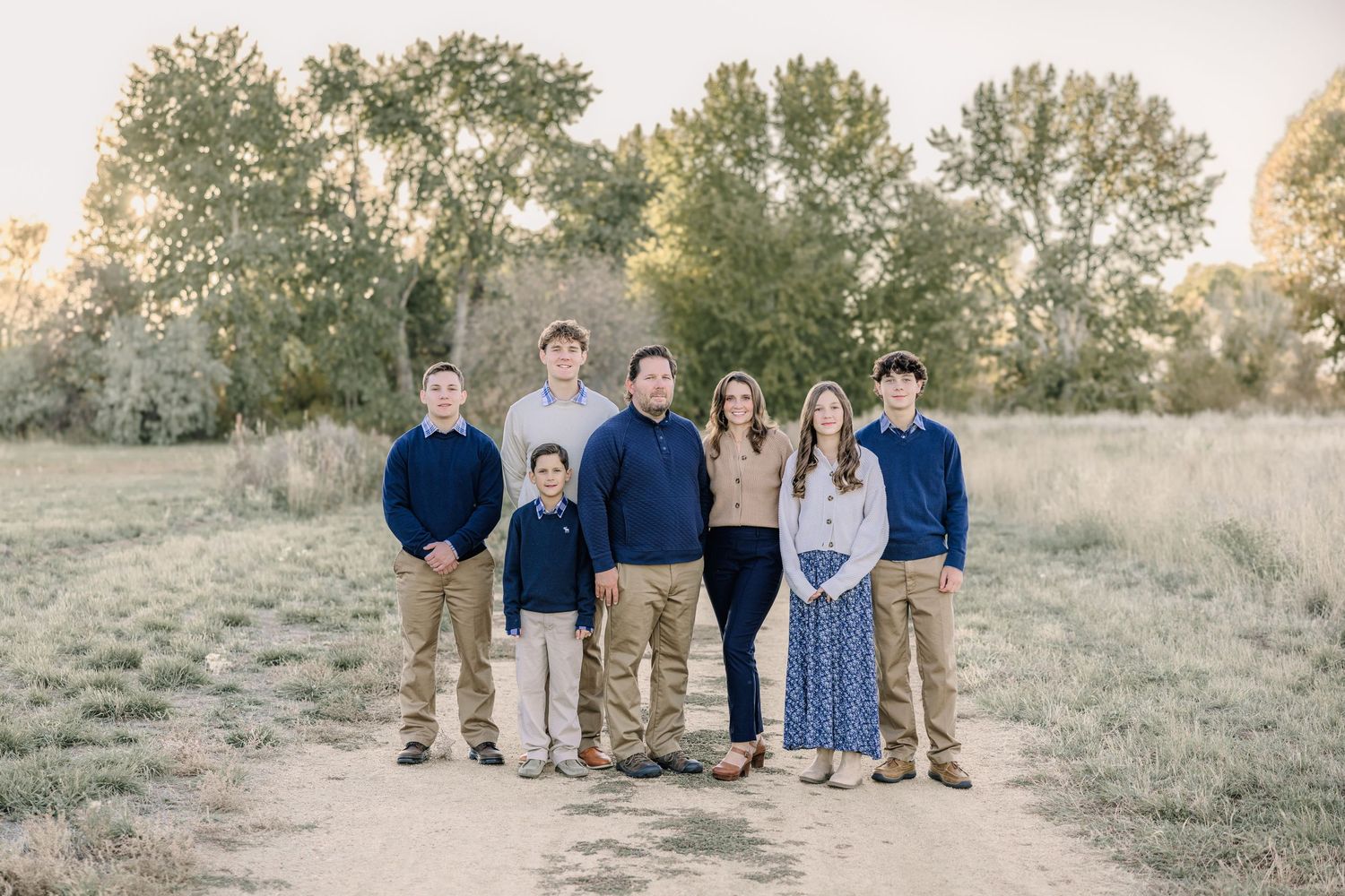 Family in coordinating blue and neutral outfits poses together in a golden grassy field at sunset for portraits by a Helena photographer