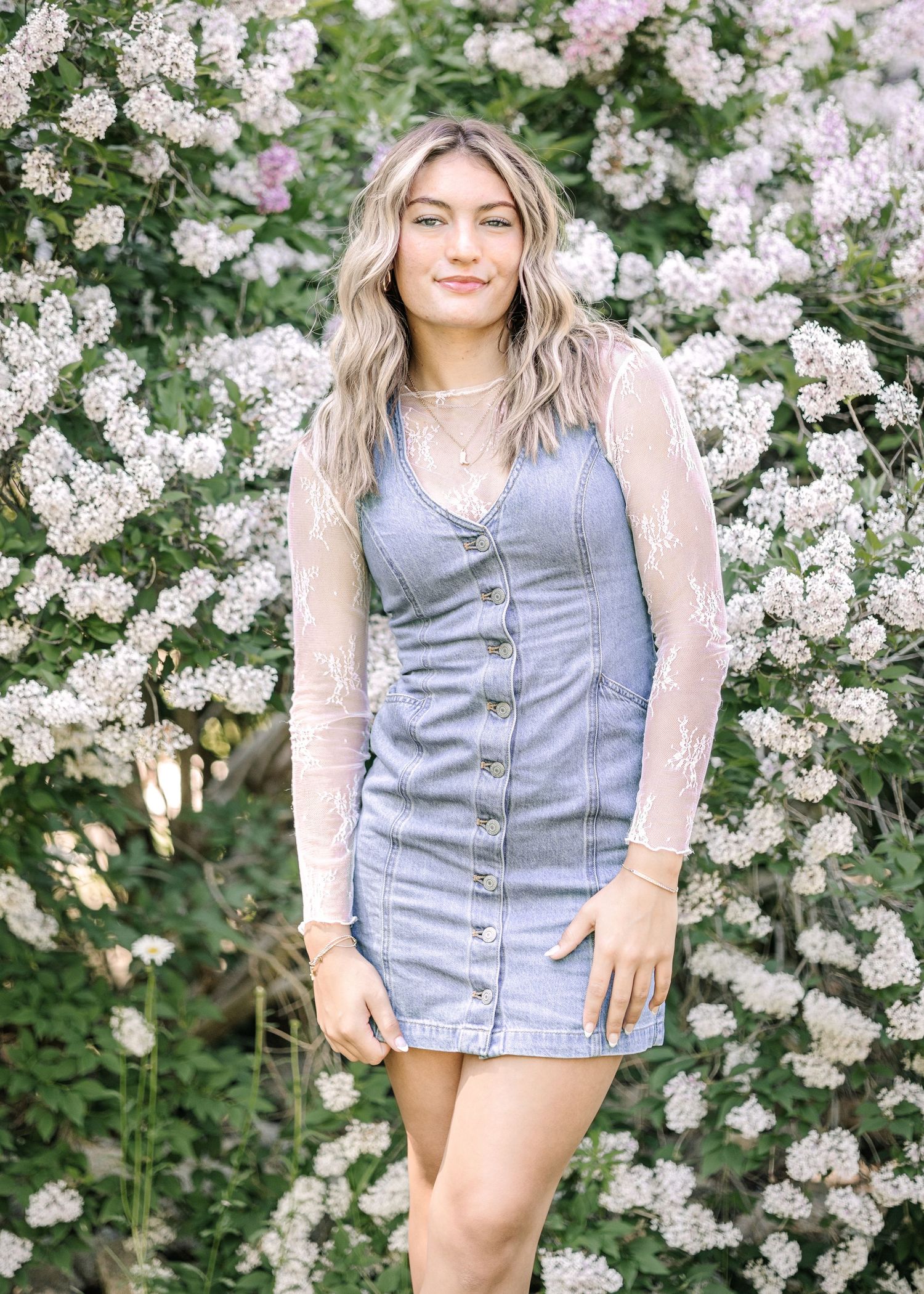Young woman in denim dress and lace top posing in front of blooming white flowering shrubs outdoors.