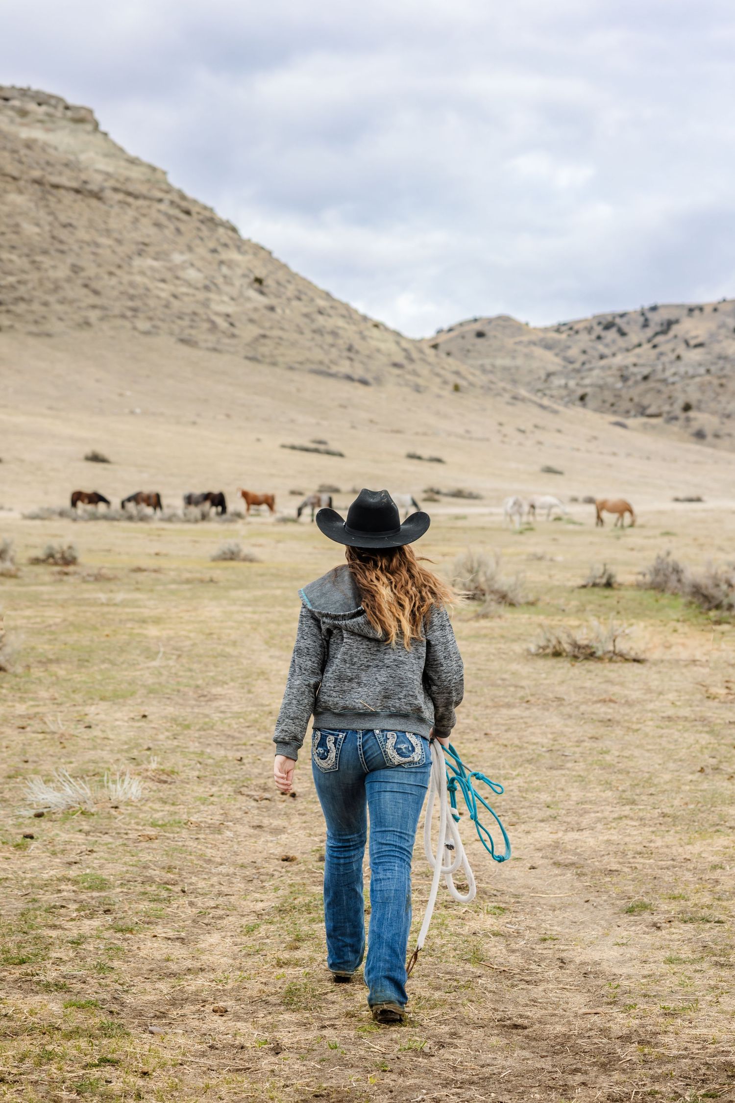 A cowgirl walks across a rugged mountain landscape with horses grazing in the distance. Photo by Montana photographer, Kelsea Laine.