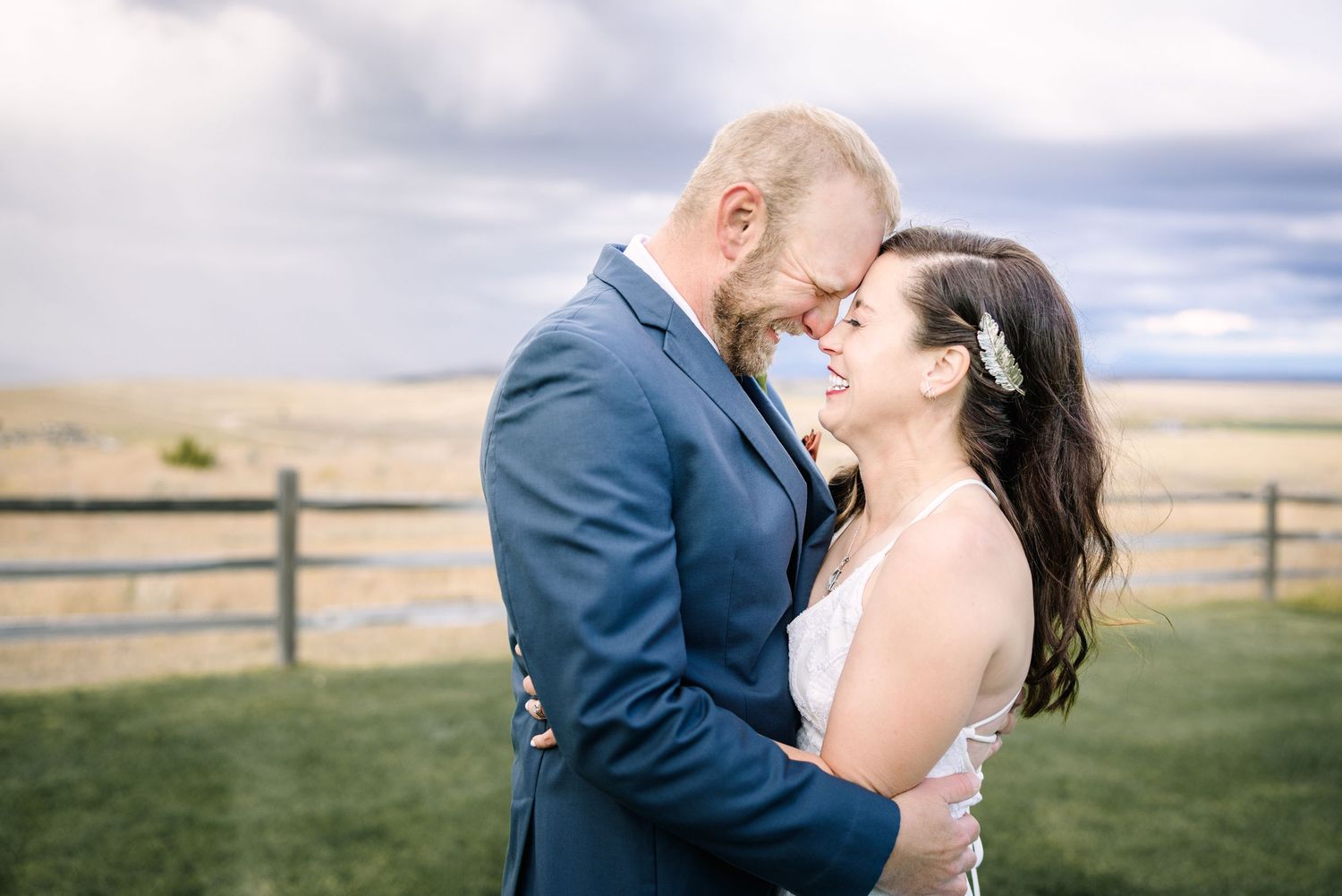 A bride and groom share a romantic forehead touch outdoors, with stormy skies and a rustic fence backdrop.