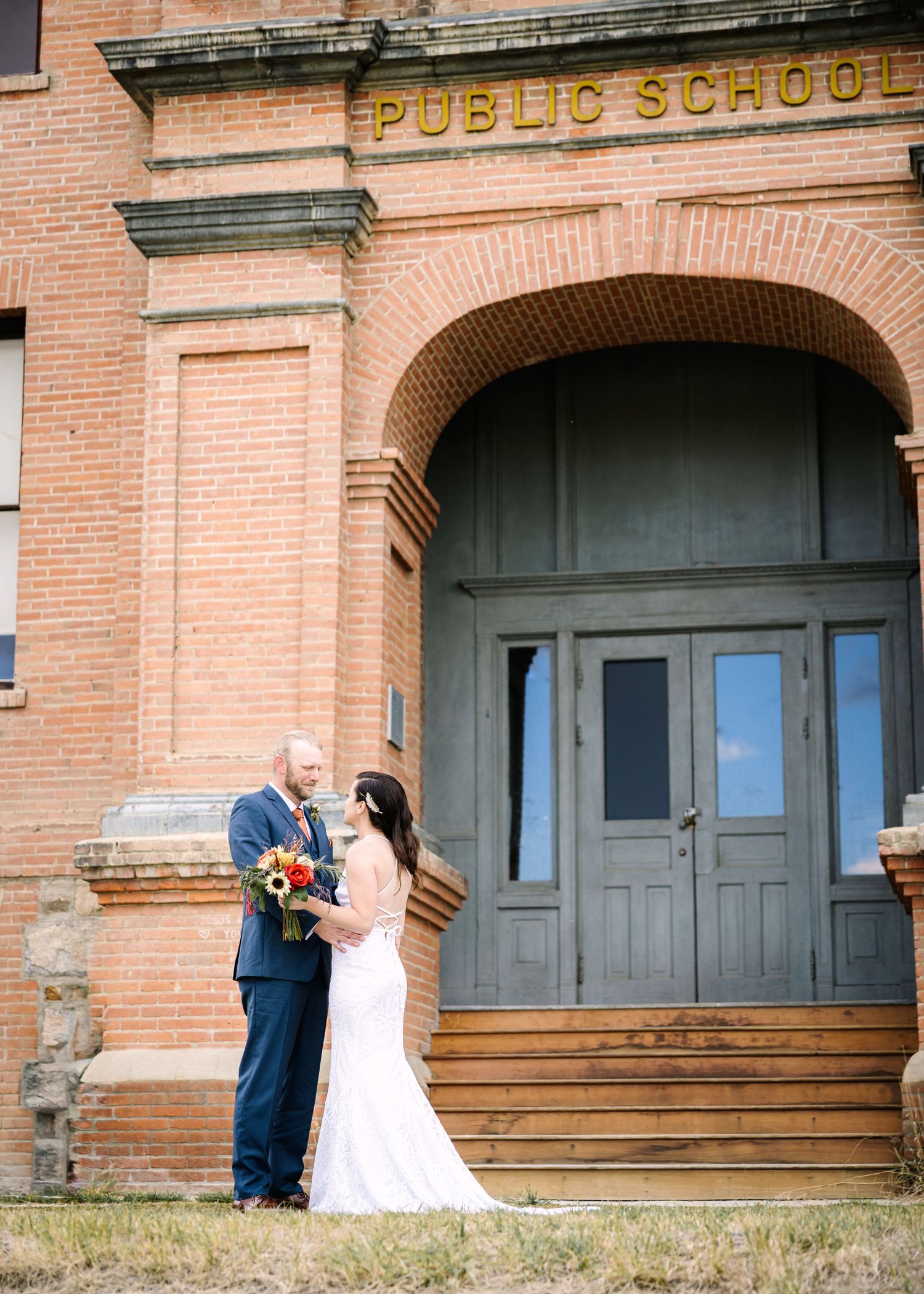 A couple shares a romantic moment in front of a historic brick public school building during their wedding photoshoot.