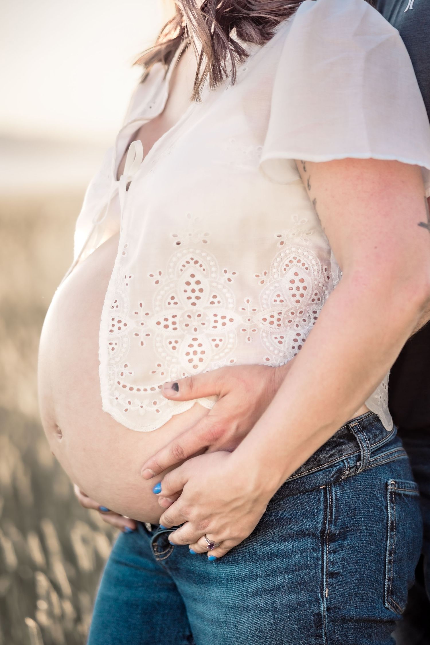 Side view of pregnant belly in white eyelet top and jeans against sunset light. A maternity photo by Kelsea Laine Photography