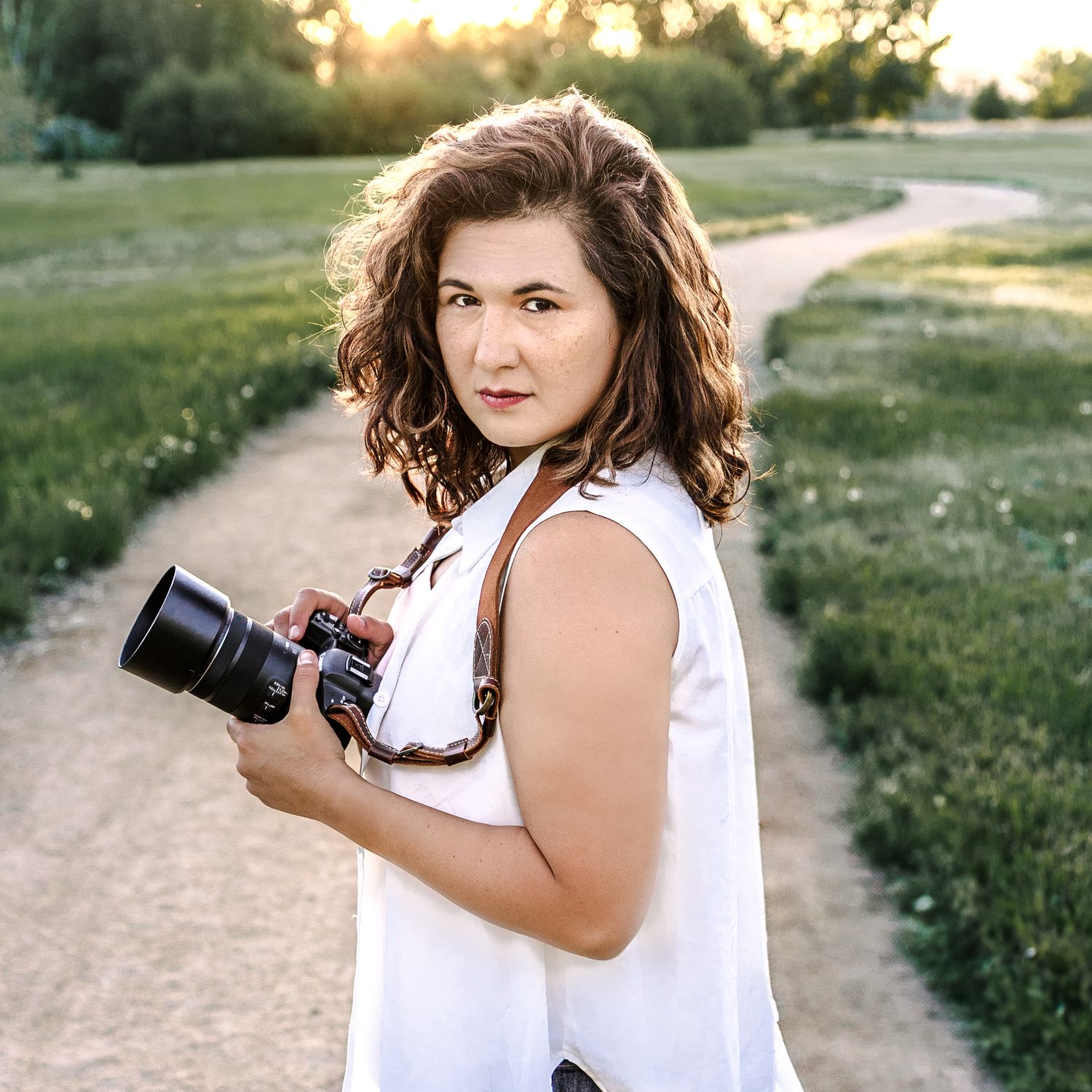 Helena Wedding Photographer in white blouse holding camera on Montana dirt path during golden hour sunset.