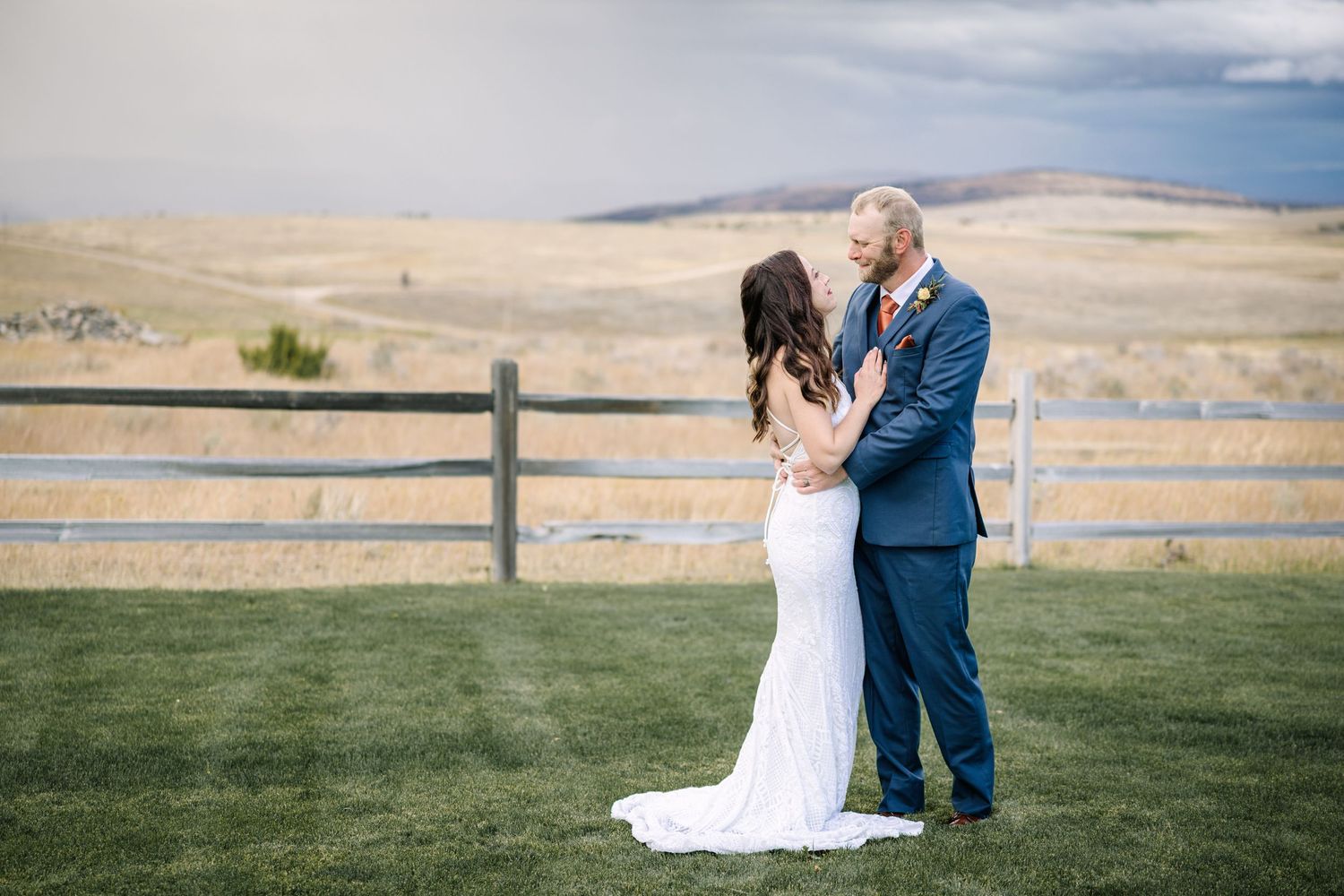 Couple in wedding attire embraces on scenic hillside ranch with wooden fence and rolling mountains in background.