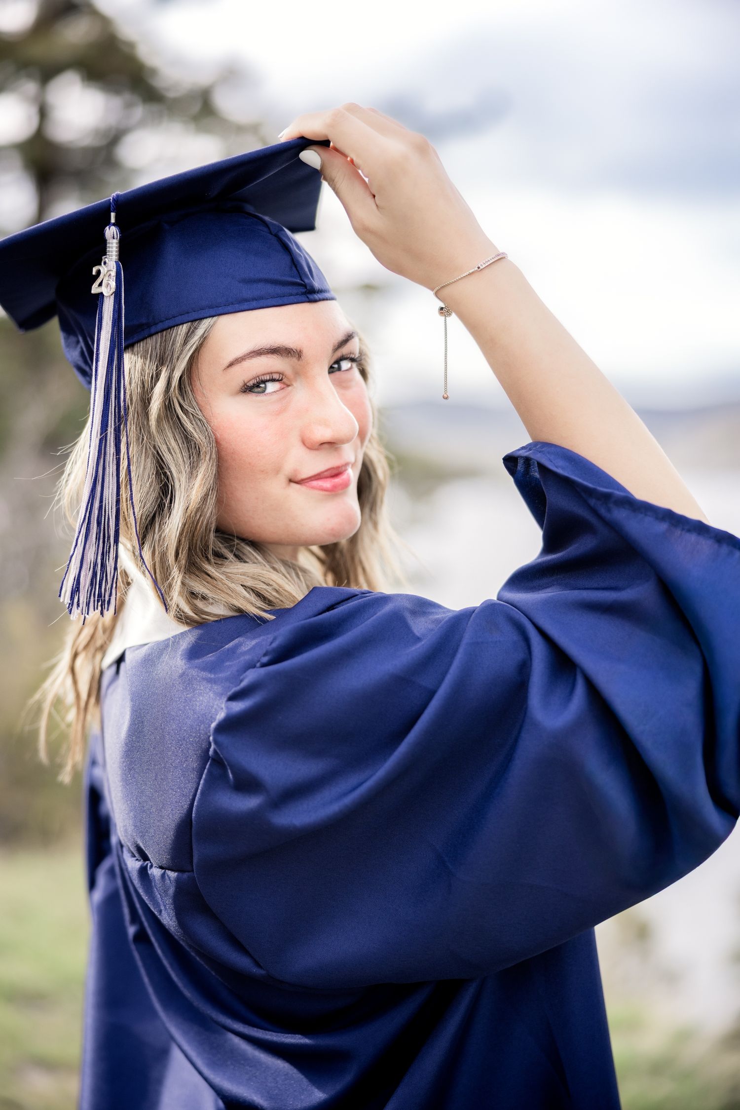 Young woman in navy blue graduation cap and gown smiles over shoulder, adjusting her cap outdoors.