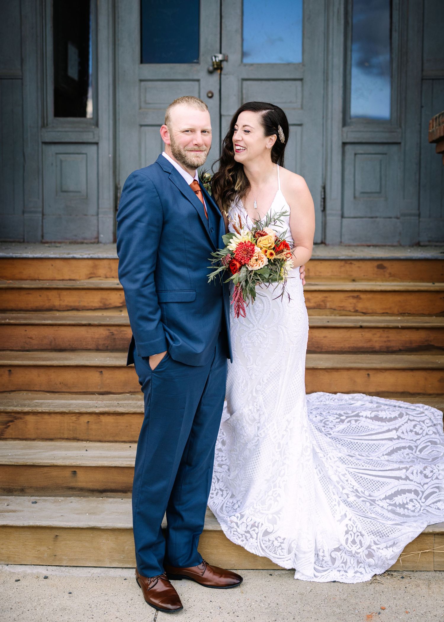 A couple shares a romantic moment on wooden steps outside a rustic building, the bride wearing a flowing lace gown.