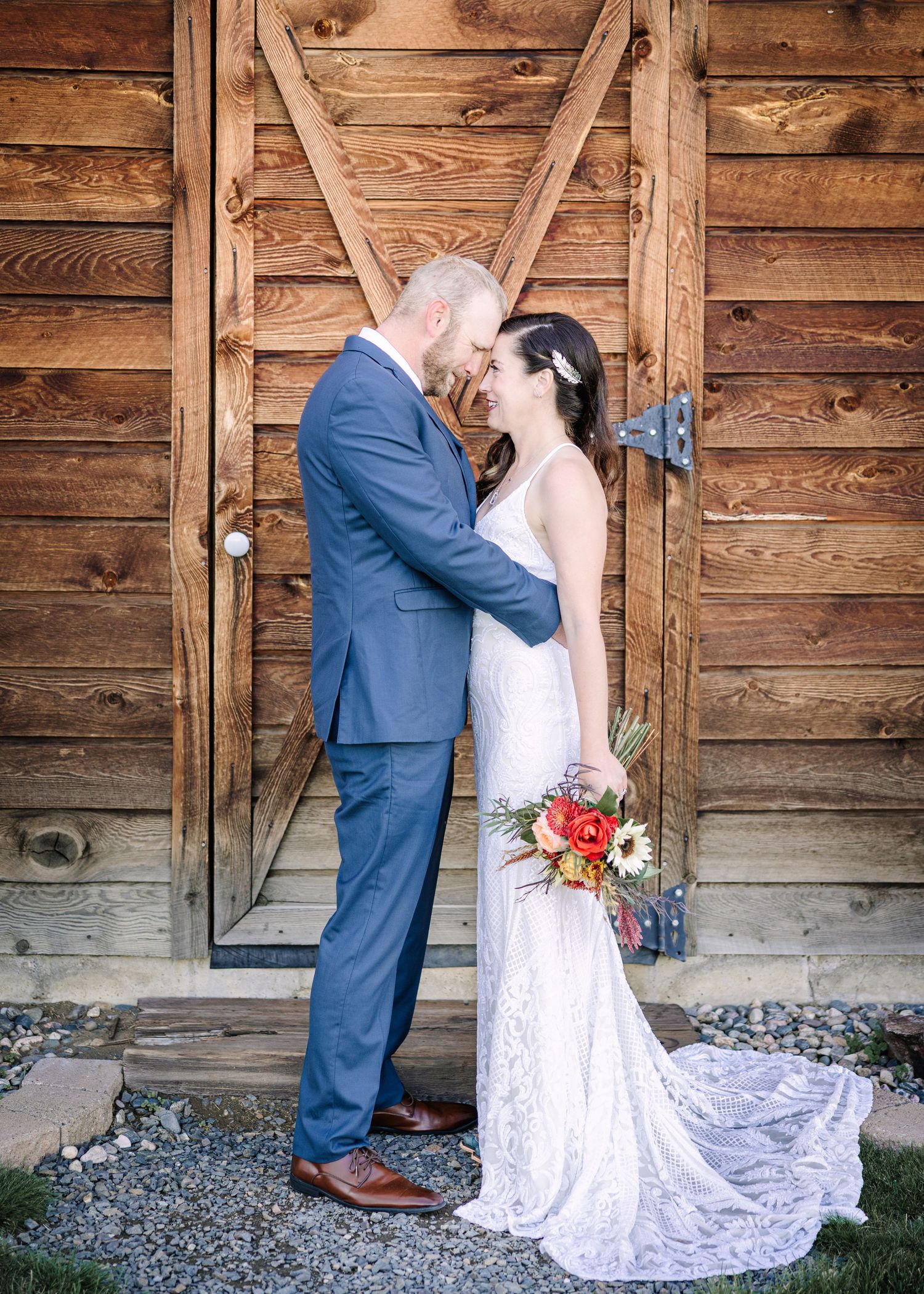 A couple shares a romantic kiss in front of a rustic wooden barn wall with a white lace dress and navy suit.