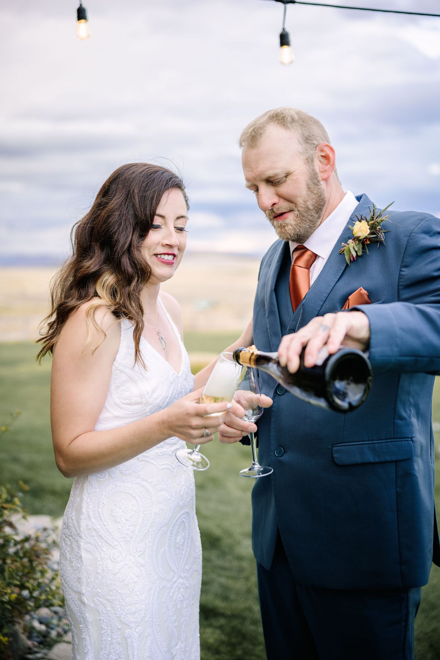 A couple laughs and shares champagne together during a wedding celebration in an outdoor setting at sunset.