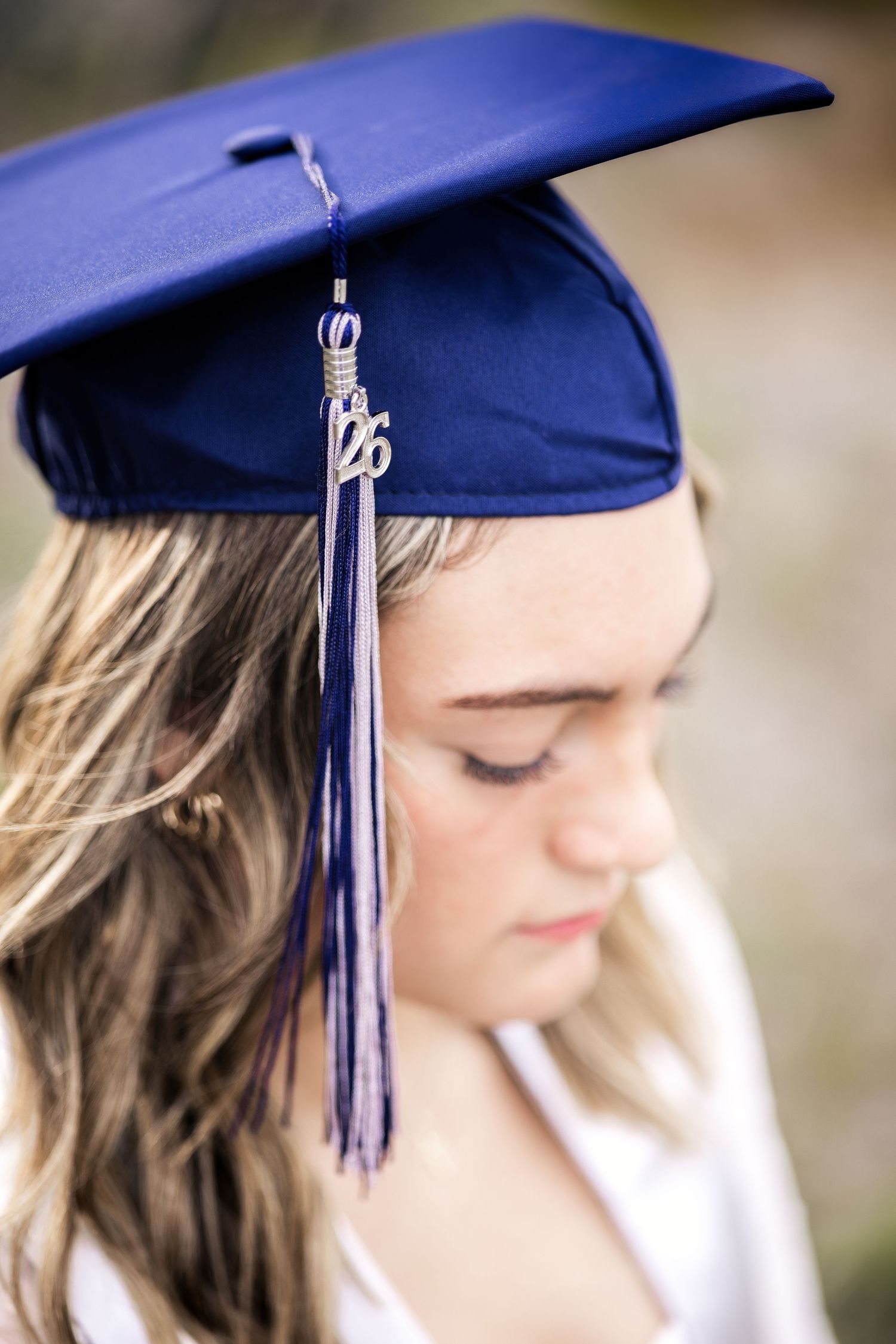Young woman wearing a navy blue graduation cap with tassel, looking down in a reflective moment outdoors.