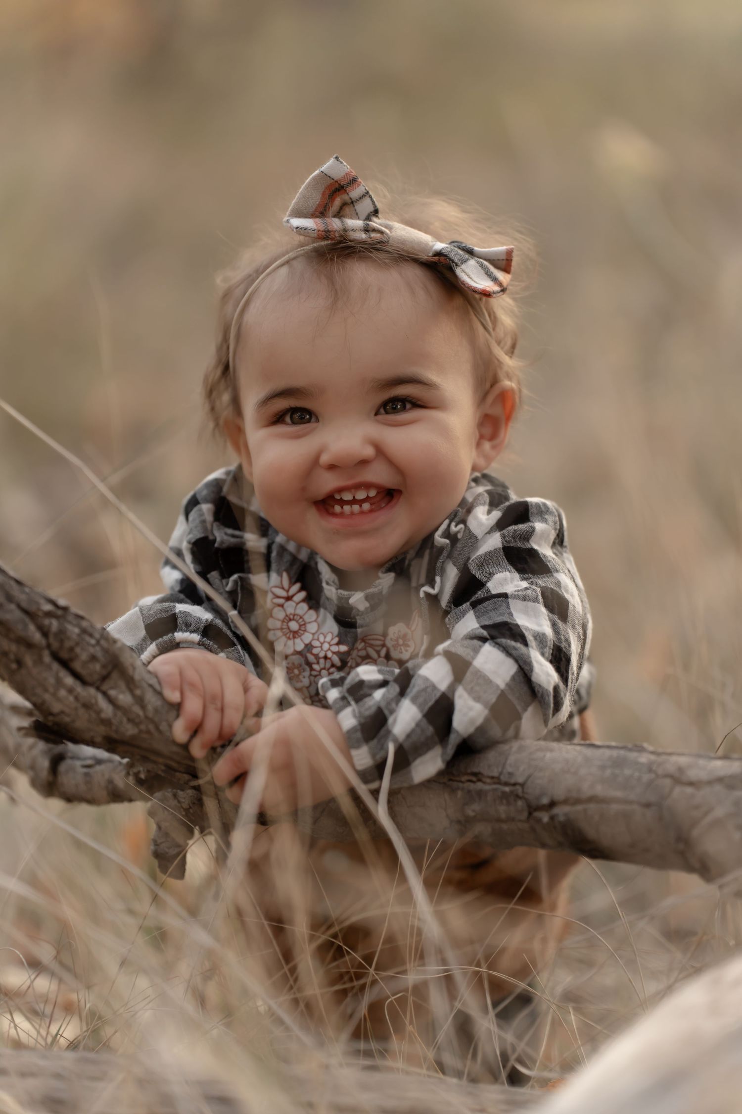 A child in black and white plaid smiles while leaning on a fallen tree branch.