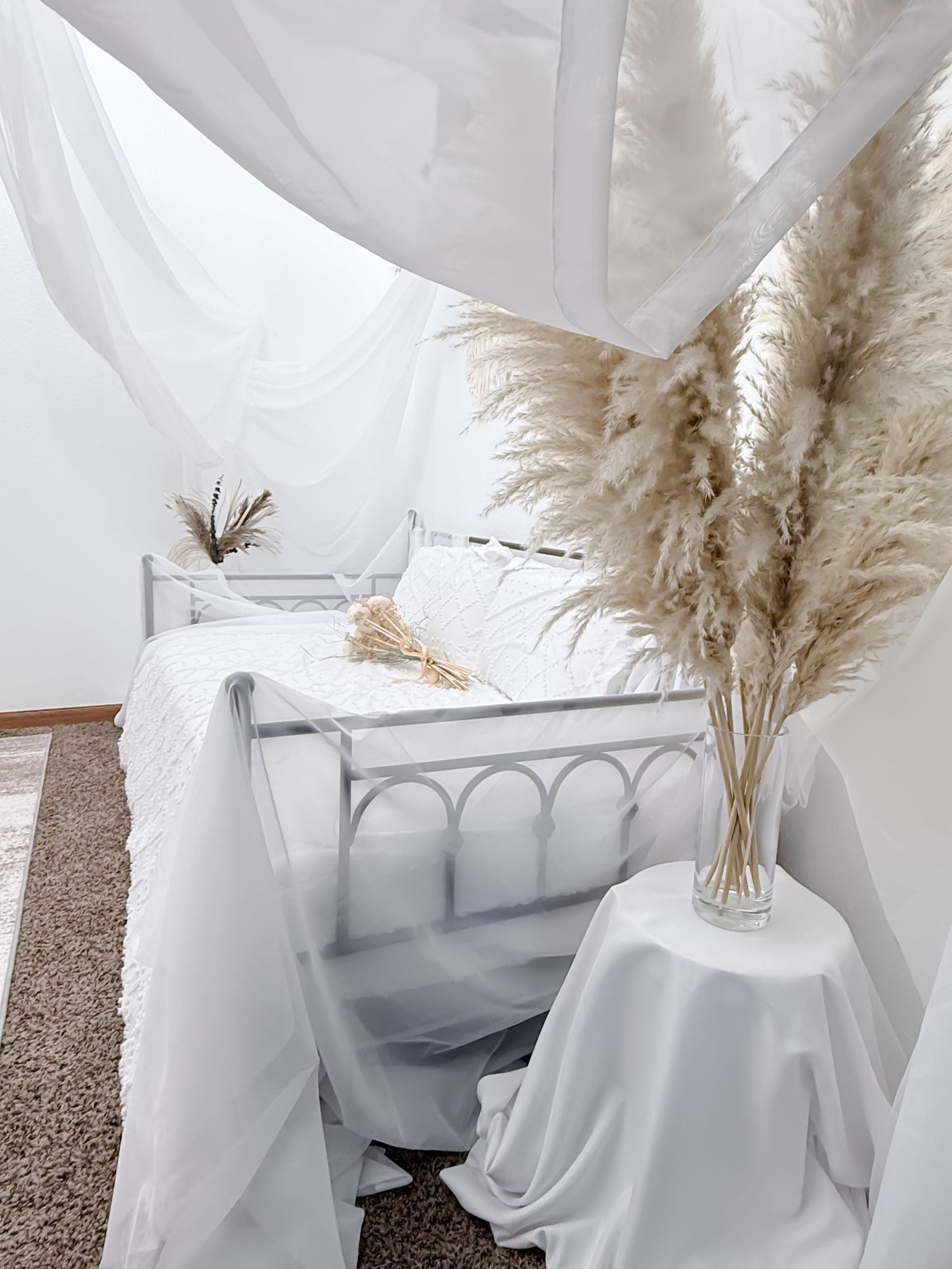 Elegant white floral arrangement featuring pampas grass in a clear vase against a soft white draped fabric backdrop.