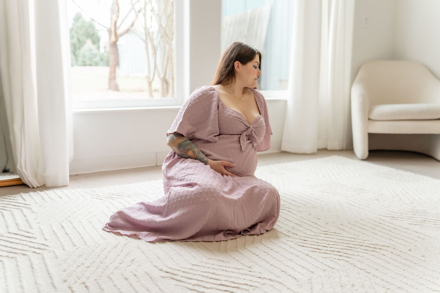 A person in a flowing dusty rose dress sits gracefully on a light carpet in a bright, airy room with white curtains.