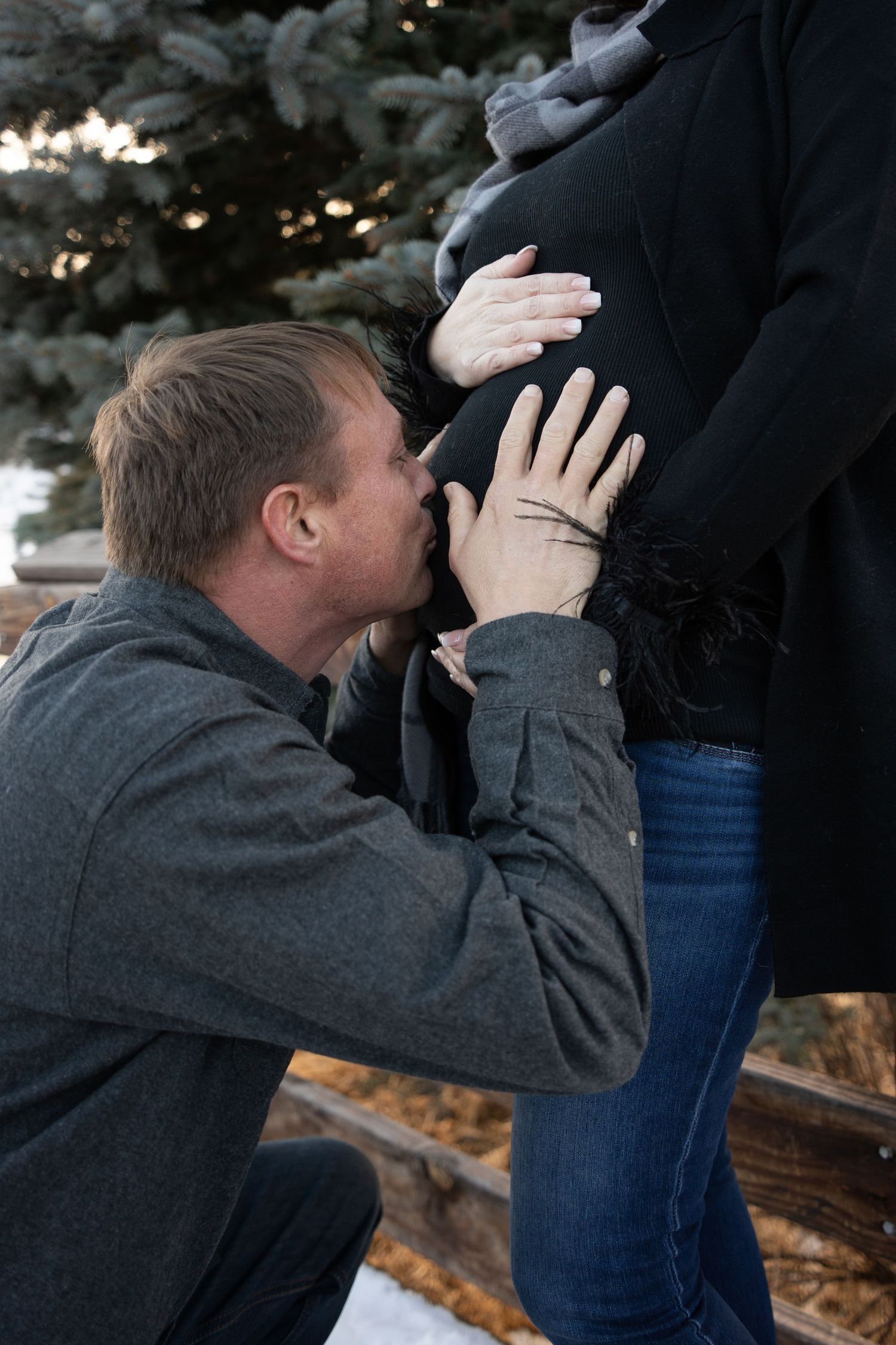 An intimate maternity moment captured in front of snow-covered evergreen trees.