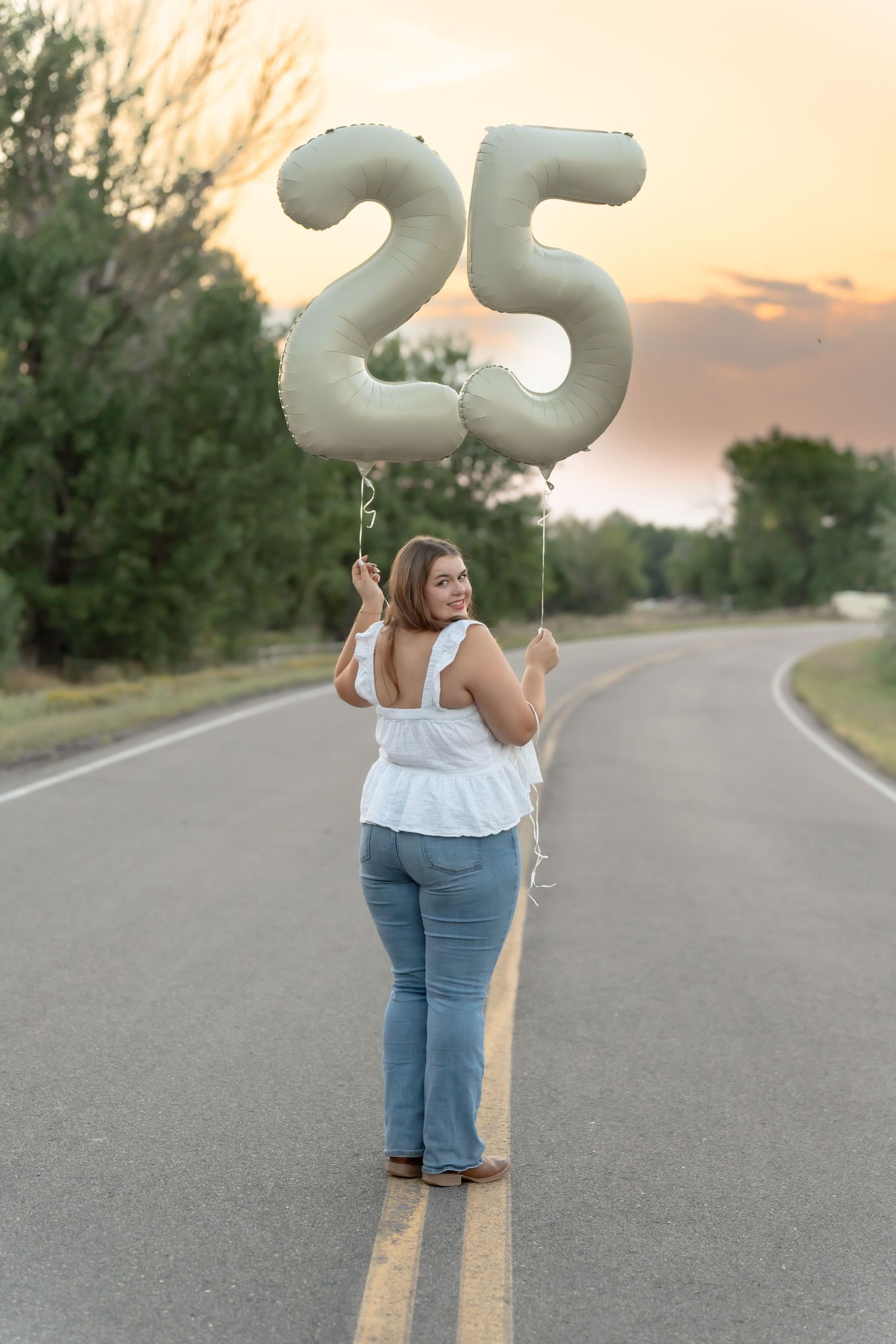 A person holding silver number balloons on an empty road at sunset.