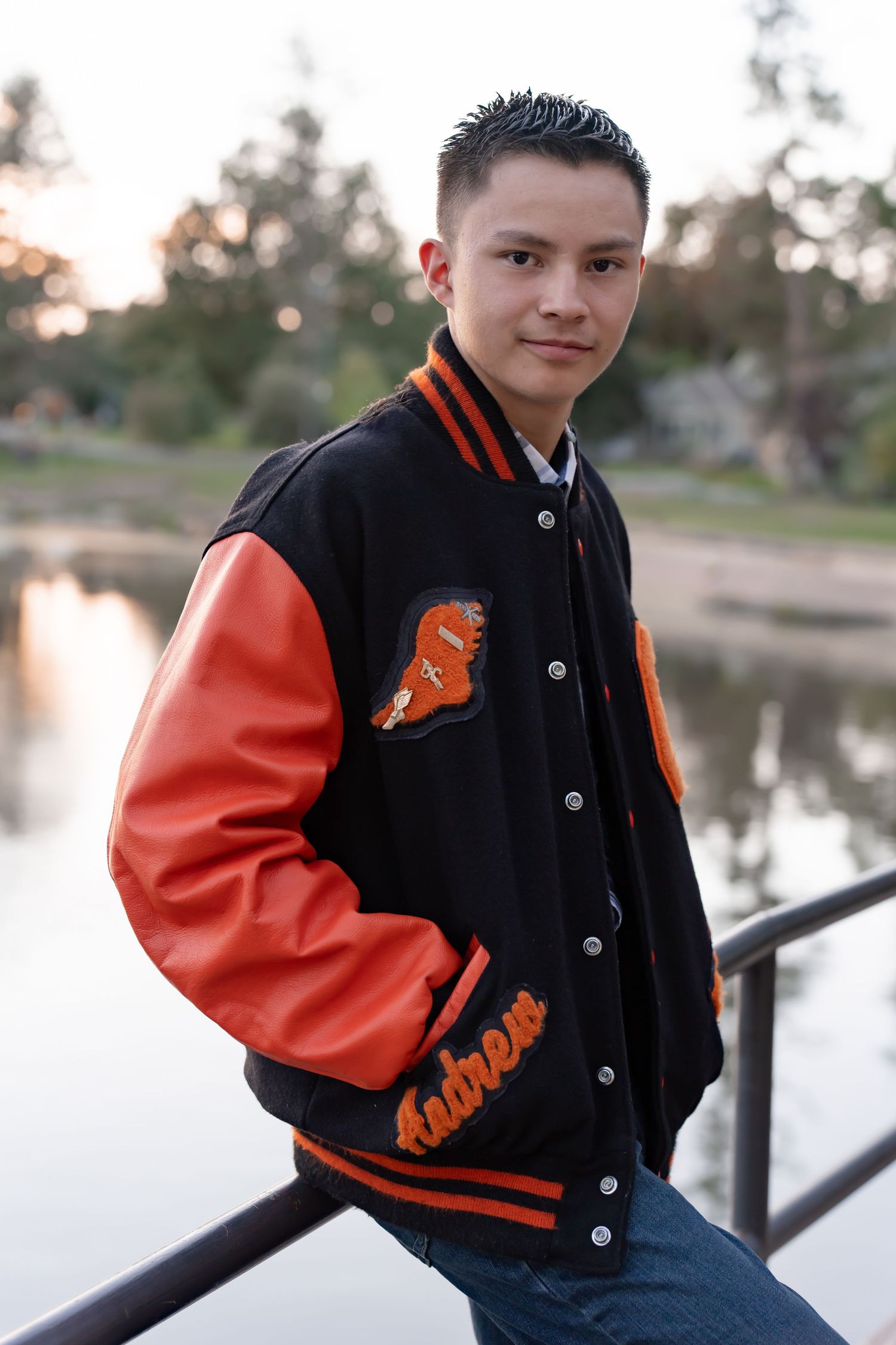 Student wearing black and orange varsity letterman jacket poses against lake backdrop during sunset.