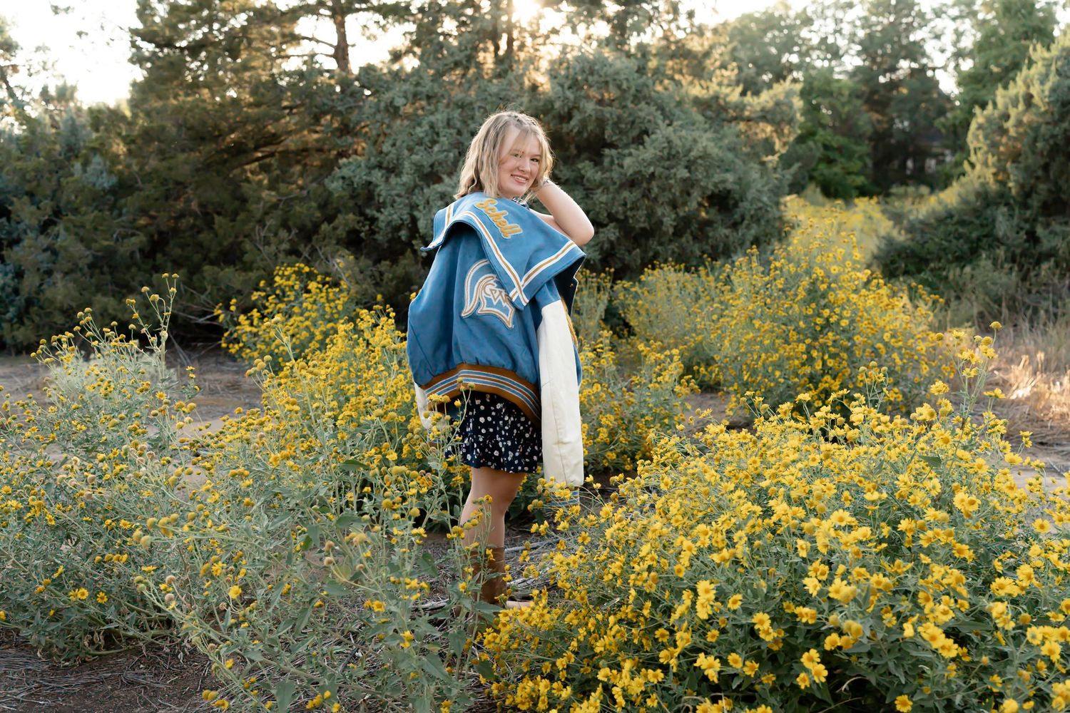A person stands among bright yellow wildflowers wearing a blue sweater and polka dot skirt in a natural outdoor setting.