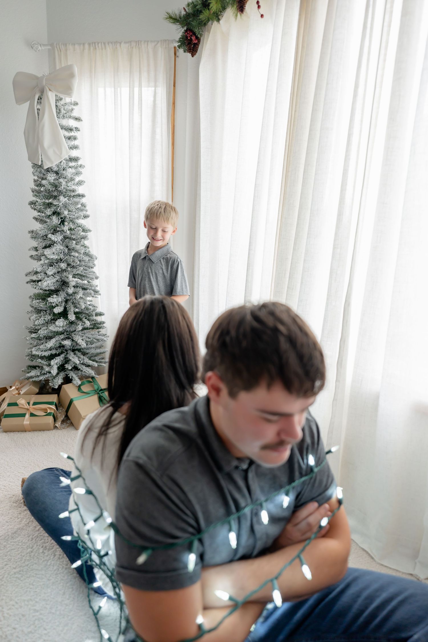 Person in gray hoodie sits near white curtains and decorative Christmas trees in a bright interior space.