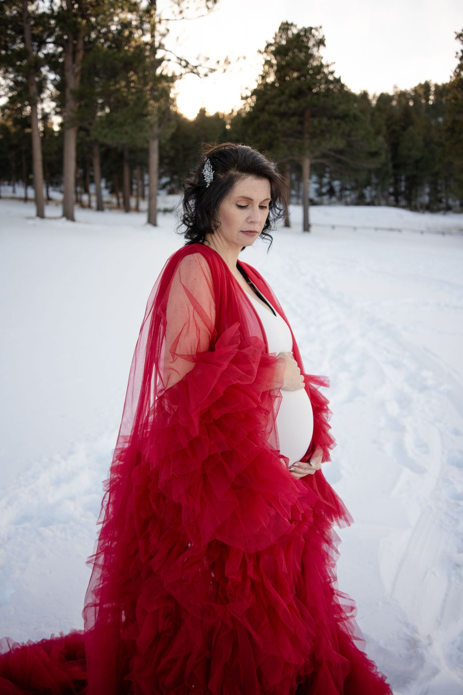 A vibrant red tulle maternity dress creates a dramatic silhouette against a snowy winter forest backdrop.