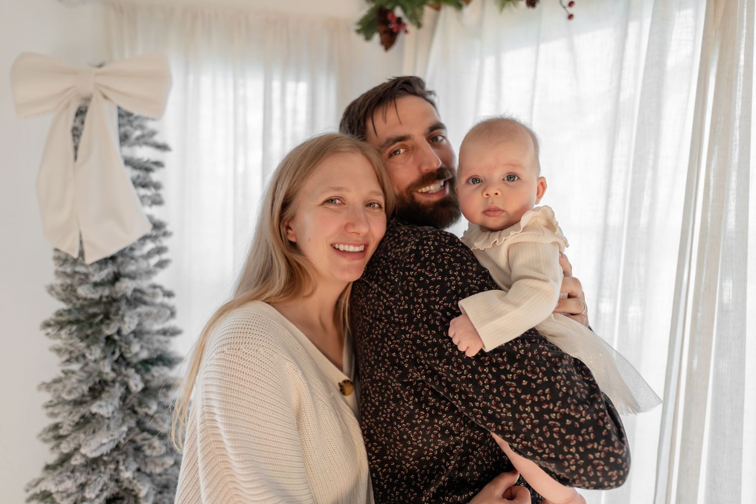 A family poses together in a bright room decorated for Christmas with a tree and white curtains in the background.