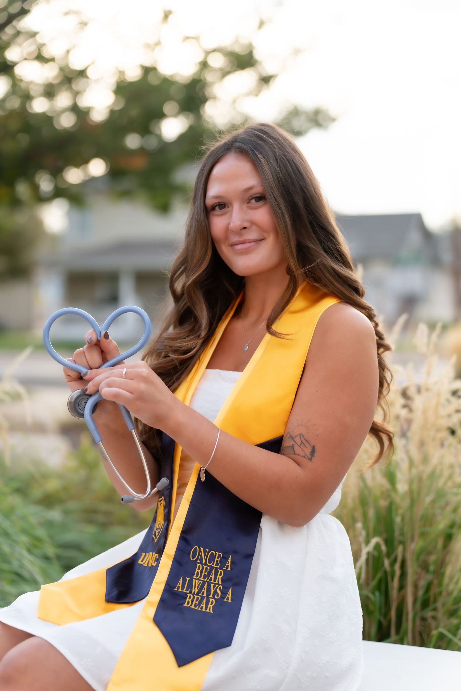 Nursing graduate in white dress and gold stole holds stethoscope in heart shape during outdoor photoshoot.