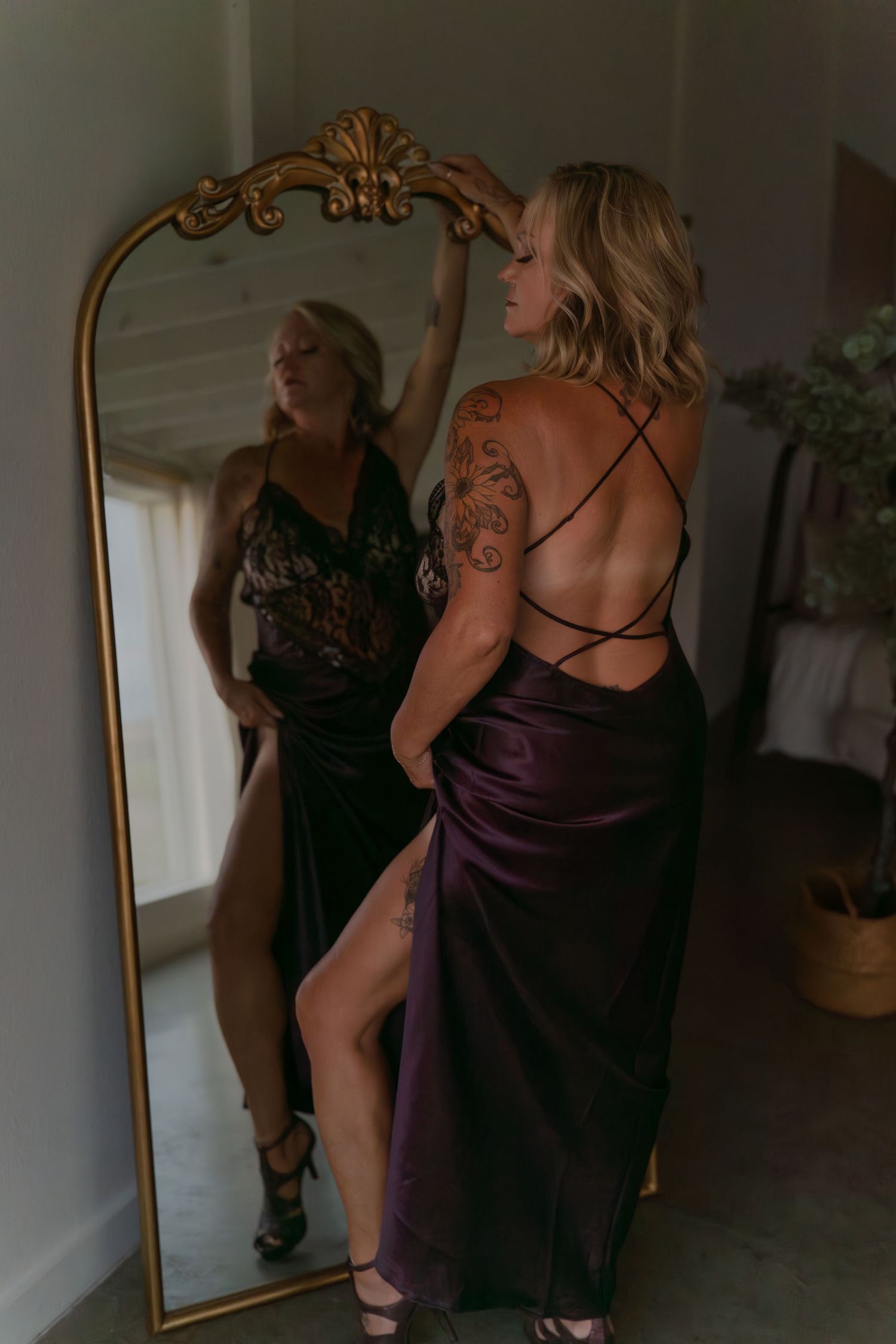 Figure in purple silk dress poses by ornate gold mirror showing reflection.