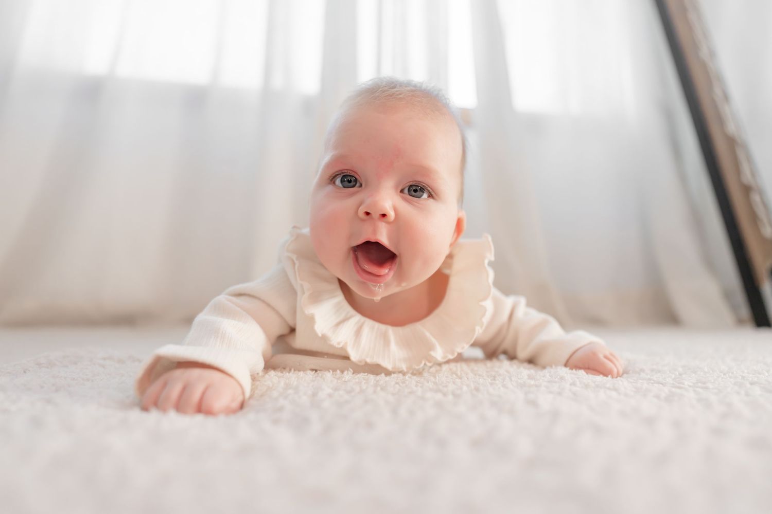 Baby in white clothing lies on a light carpet while smiling and looking up at the camera.