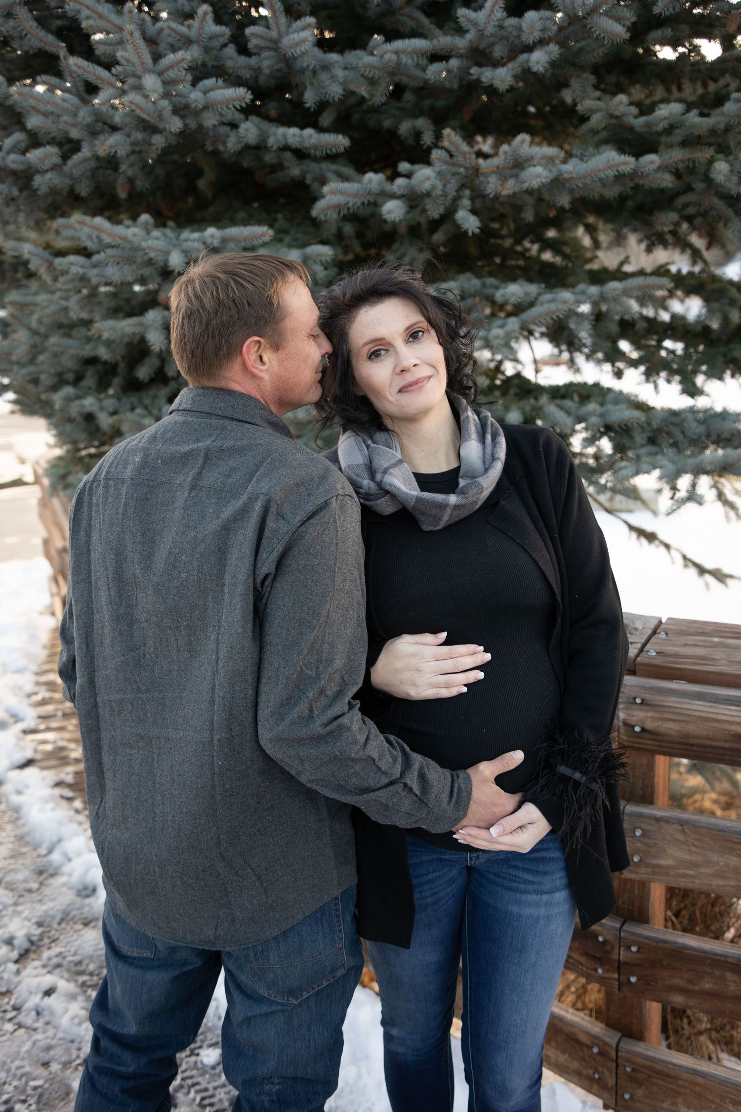 A winter maternity photo of a couple standing by snow-covered evergreen trees.