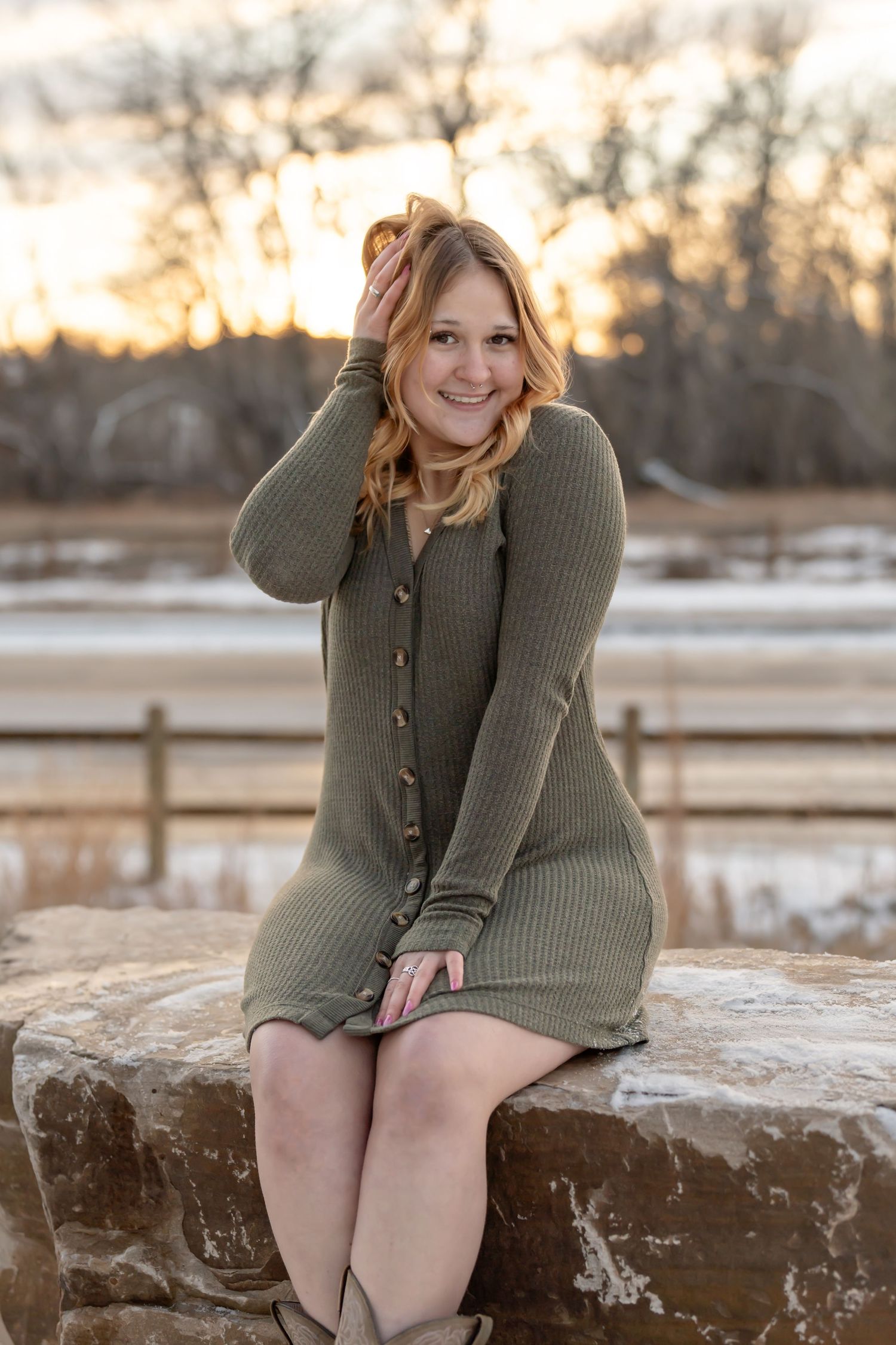 Winter portrait of person in olive sweater dress sitting on rock during sunset.