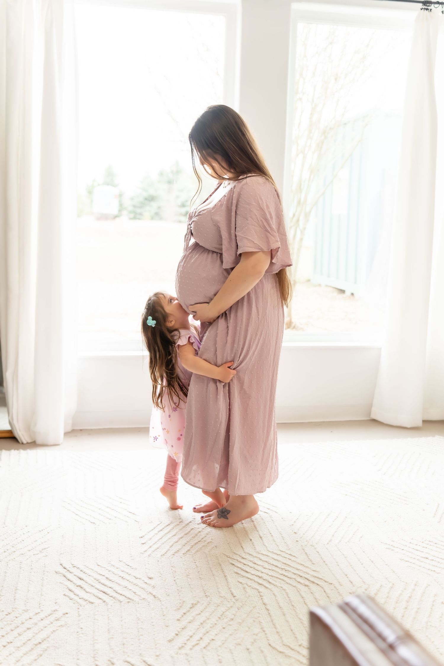 Maternity photo session in a bright white room with sunlit windows and white carpet.