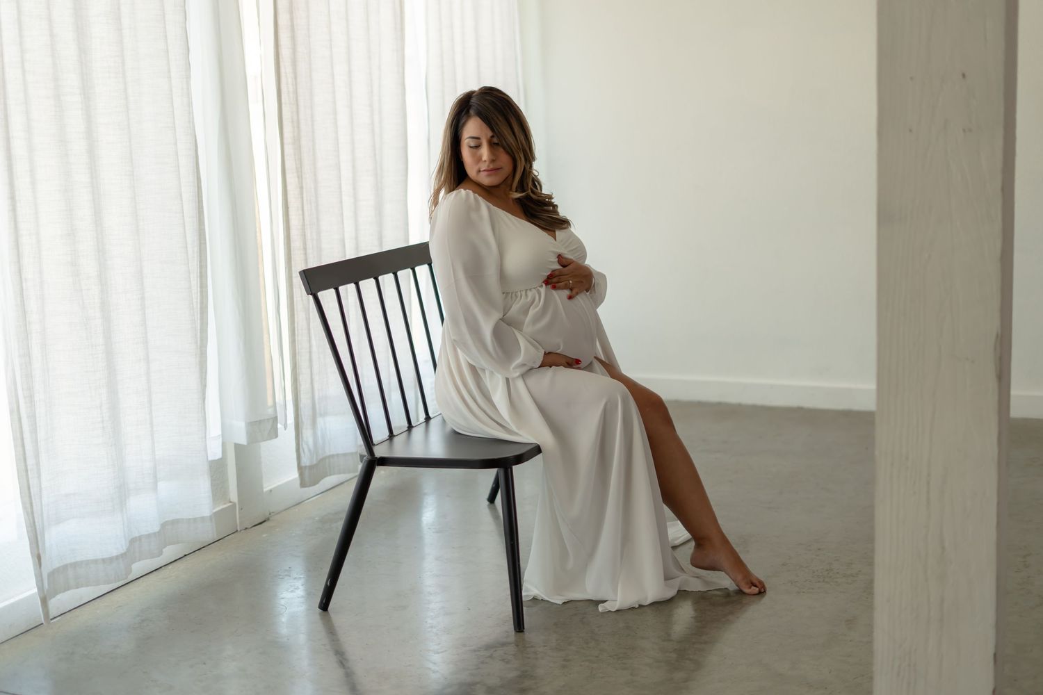 Pregnant woman in white dress posing elegantly by window in minimalist setting.