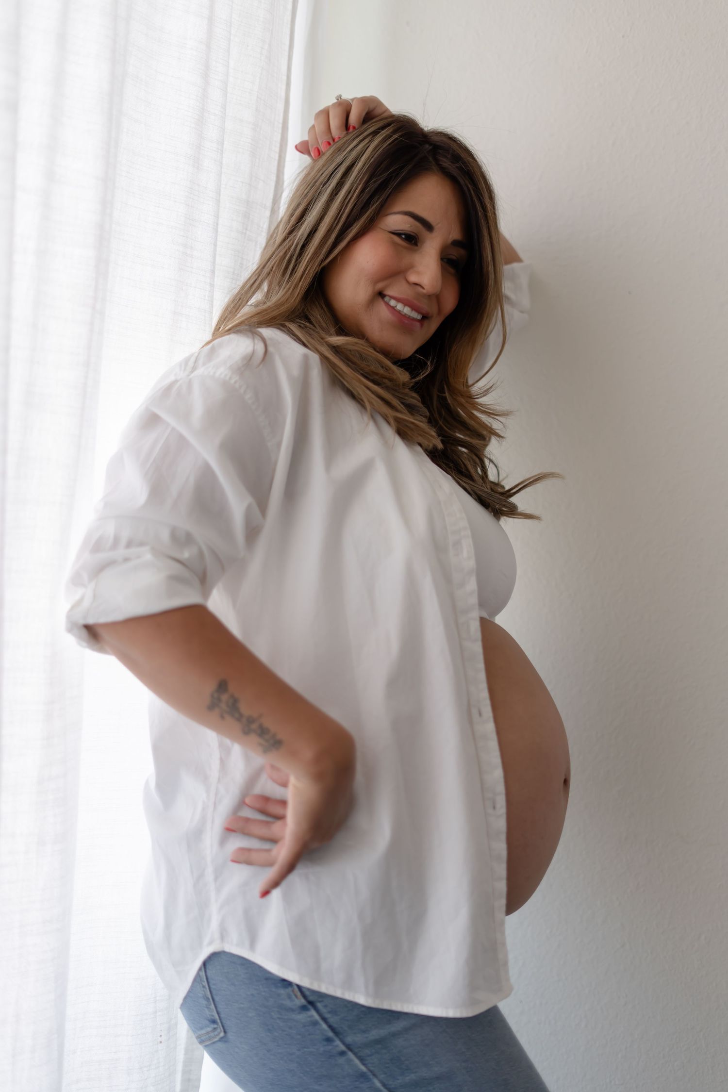 Expecting mother in white shirt and jeans poses by window during maternity photoshoot.