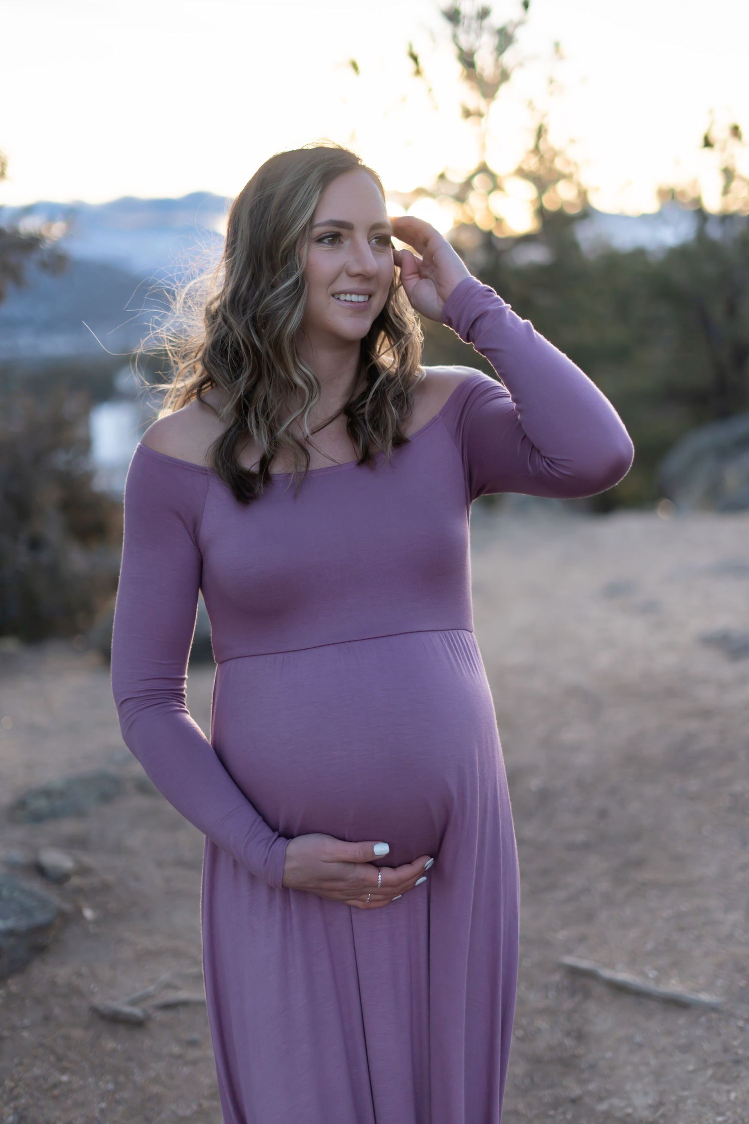 A pregnant woman in a purple maxi dress poses thoughtfully against a mountain backdrop.