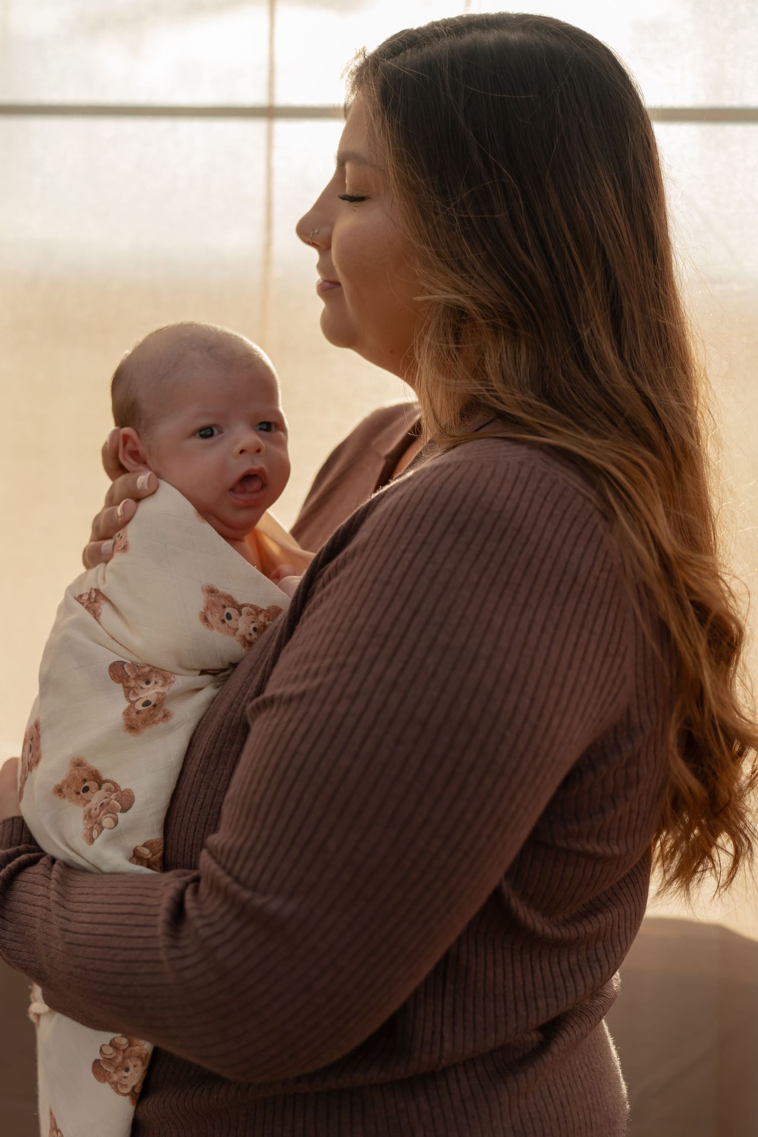 A parent in a brown sweater holds and comforts a small baby in a white onesie near a sunlit window.