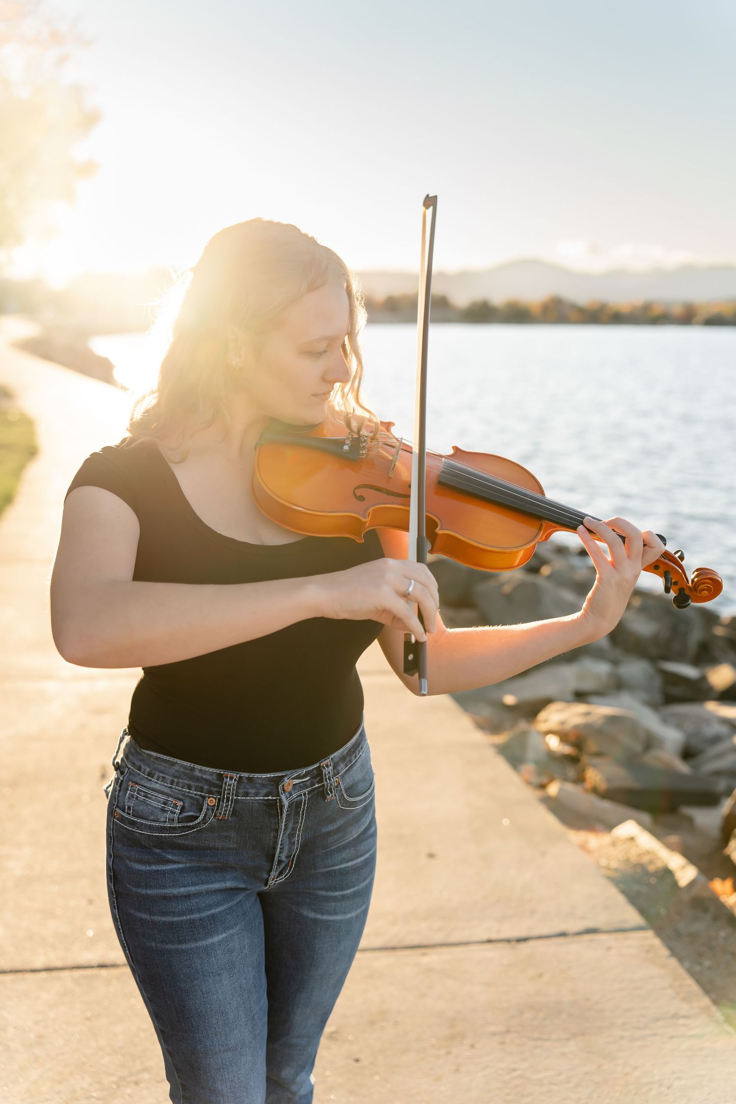 Musician playing violin outdoors by the water during golden hour sunset.