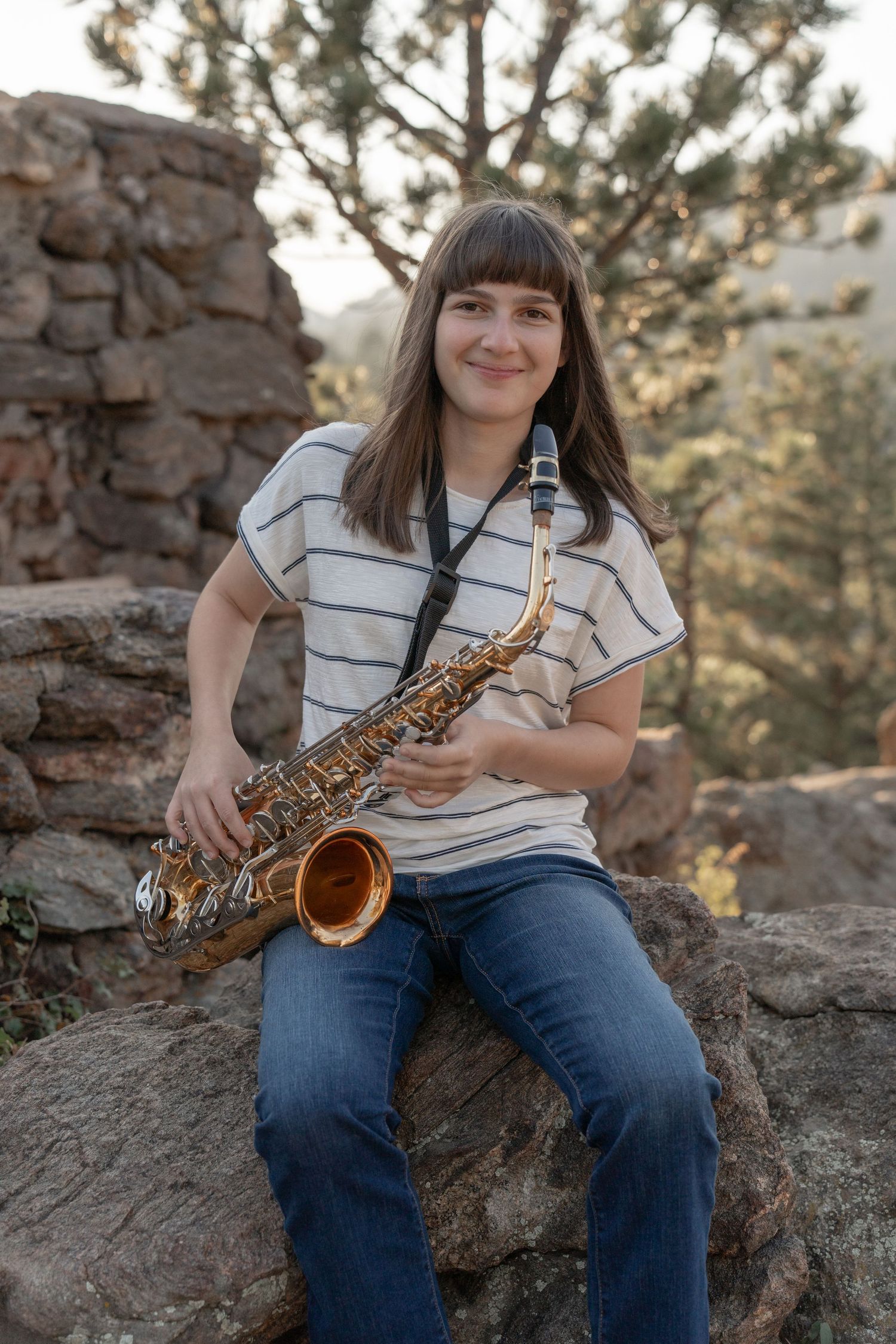 A musician poses with a saxophone on rocky terrain surrounded by pine trees.