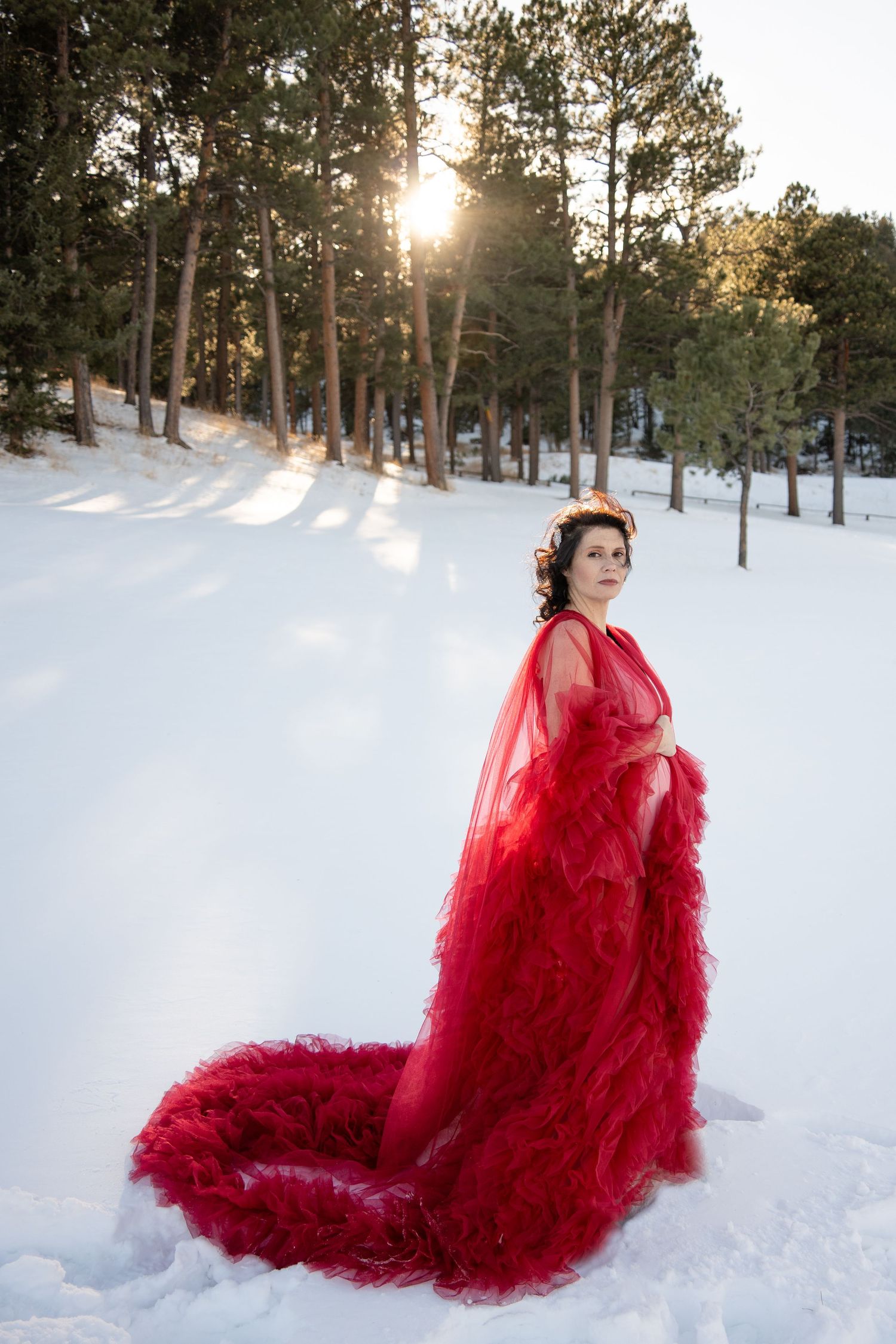 Figure in flowing red gown stands elegantly in snowy forest with sunlight streaming through trees.