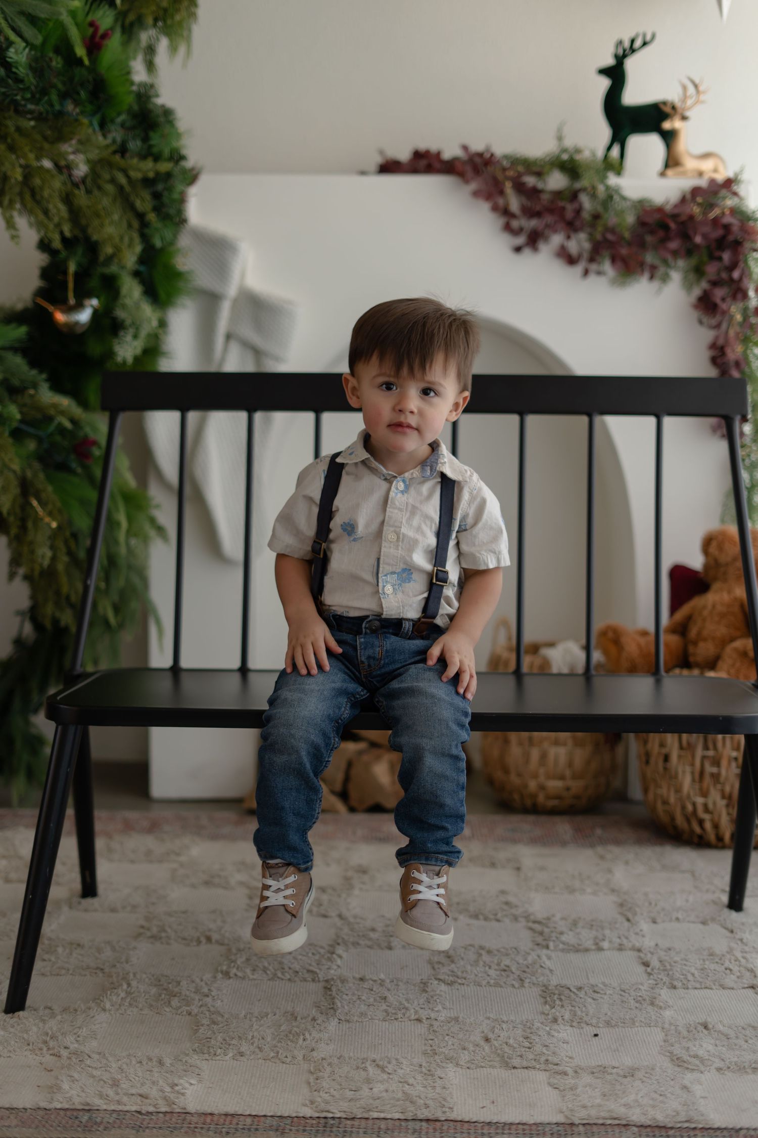 Small child wearing suspenders and jeans sits on black bench with greenery and floral decorations in background.