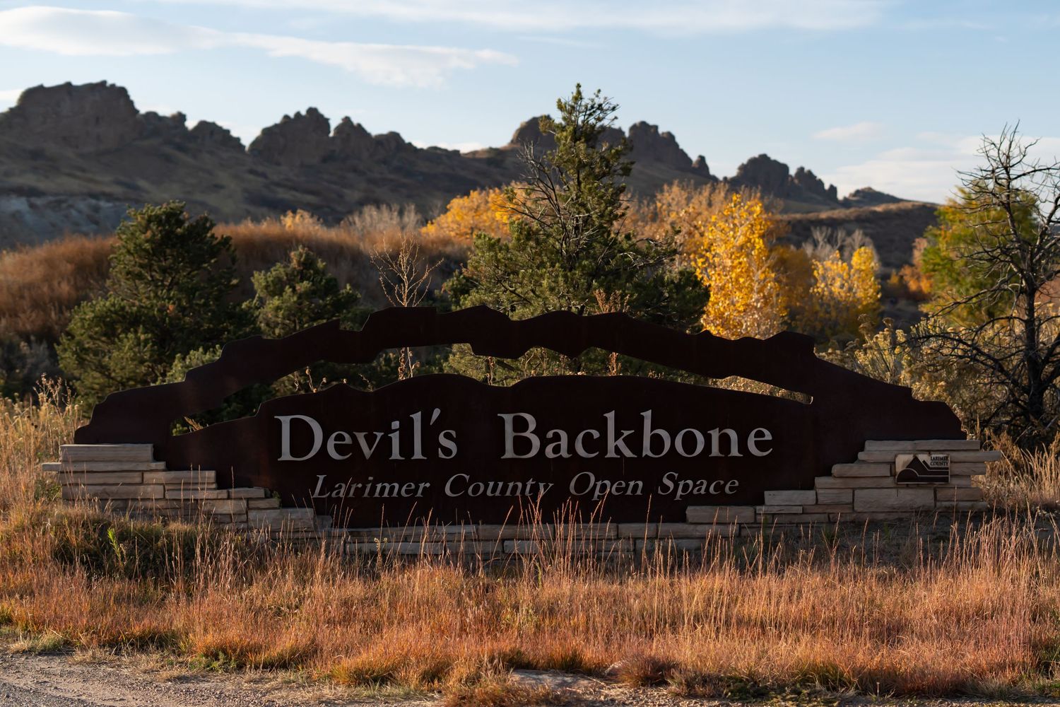 Entrance sign for Devil's Backbone Open Space in Colorado with rocky ridge and autumn foliage in background.