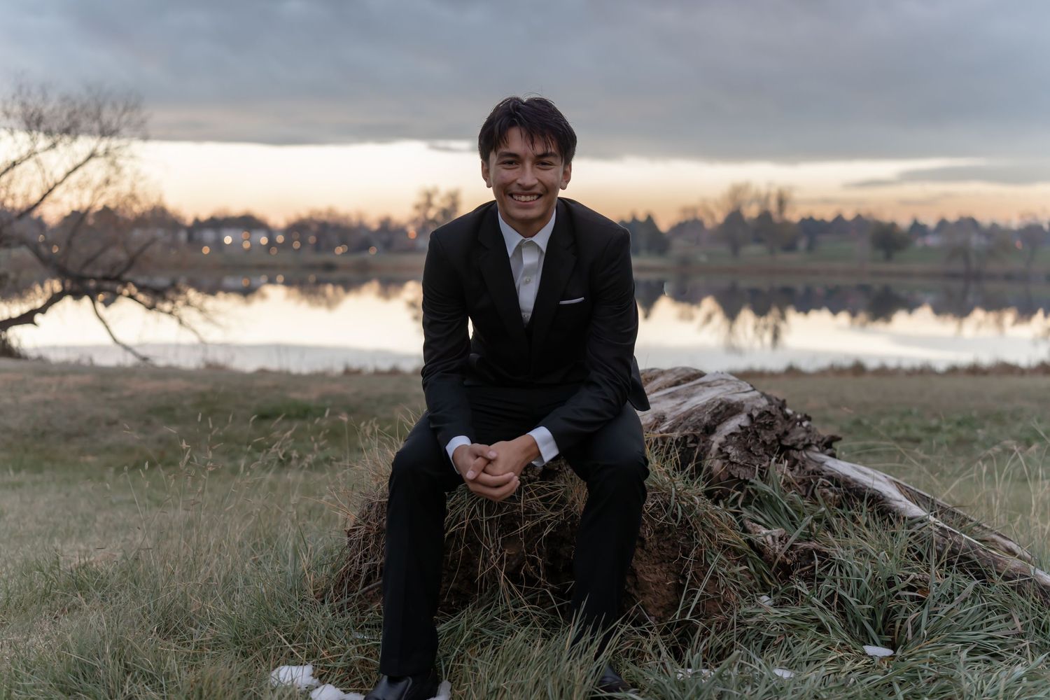 Person in black suit sits on fallen log by lake at dusk.
